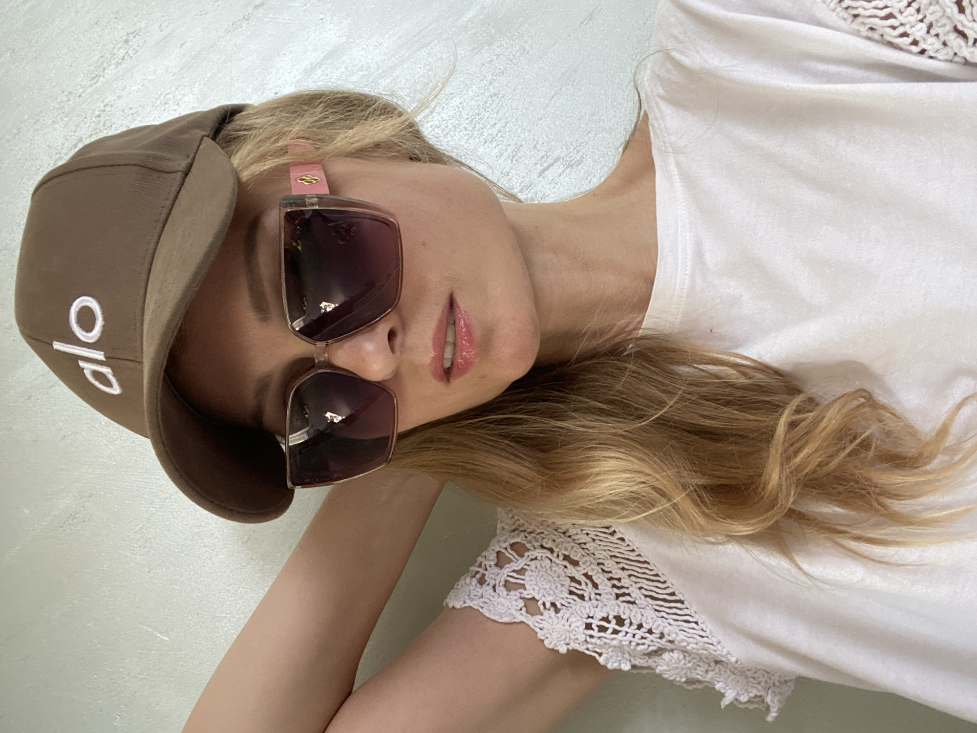 A woman with blonde hair is wearing a brown baseball cap with the logo "alo" in white letters. She also has on oversized sunglasses with pink accents and a white t-shirt with white crochet detailing on the sleeves. Her left arm is behind her head, and she is looking downwards. The background is a textured light-colored wall. This image was taken in the city of Mihanaivitski sel'ski saves, Belarus.