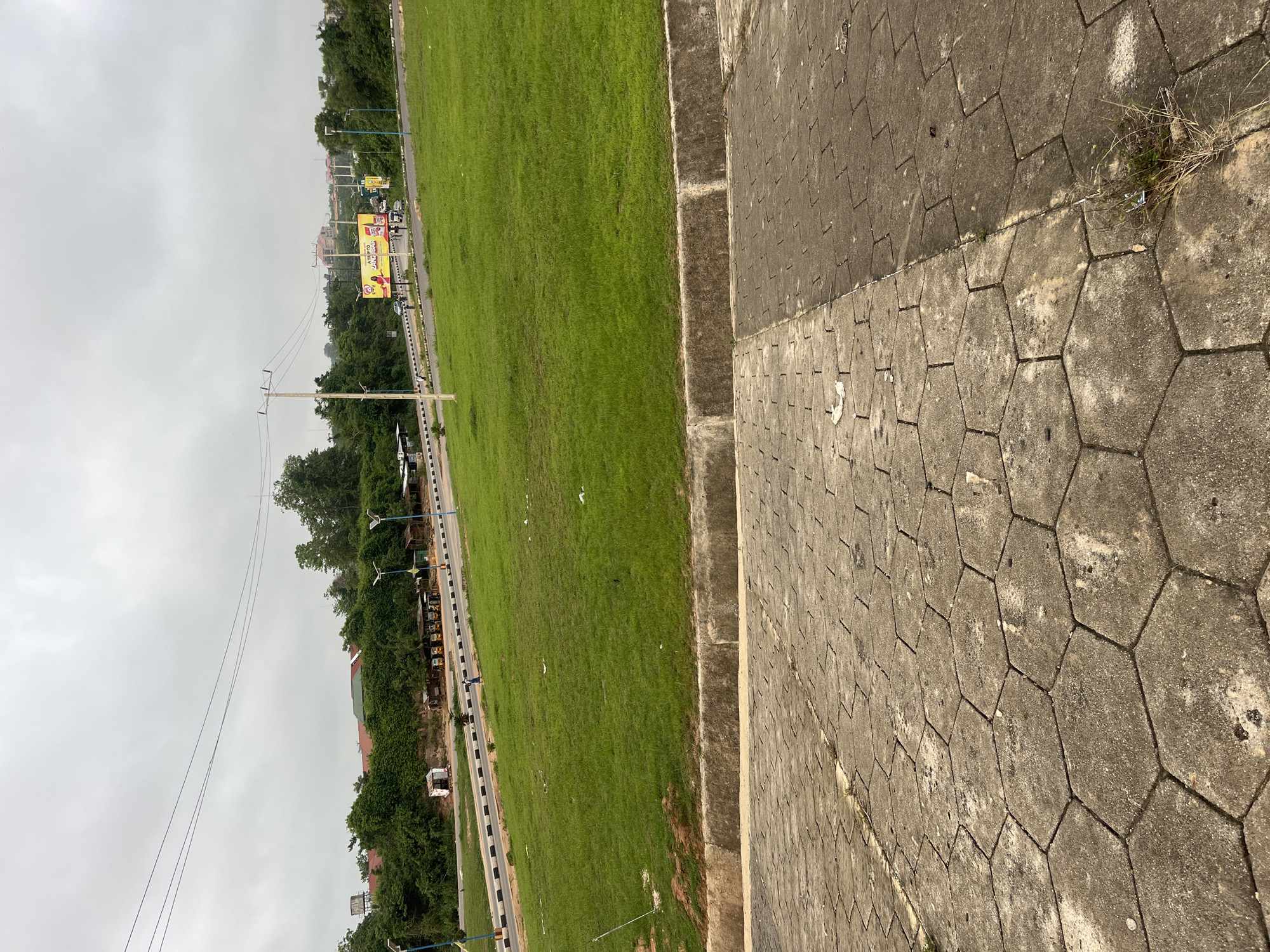 The image depicts a street in Osogbo, Nigeria, distinguished by a green lawn separating a paved walkway from a road. A prominent billboard advertising a product is visible on the other side of the road, alongside power lines and dense vegetation. The walkway is constructed of grey hexagonal pavers, some of which are stained or discolored. Overcast skies create a muted, grey light. A person walks along the road, which also contains cars, possibly in a line. A curb separates the grass from the walkway.