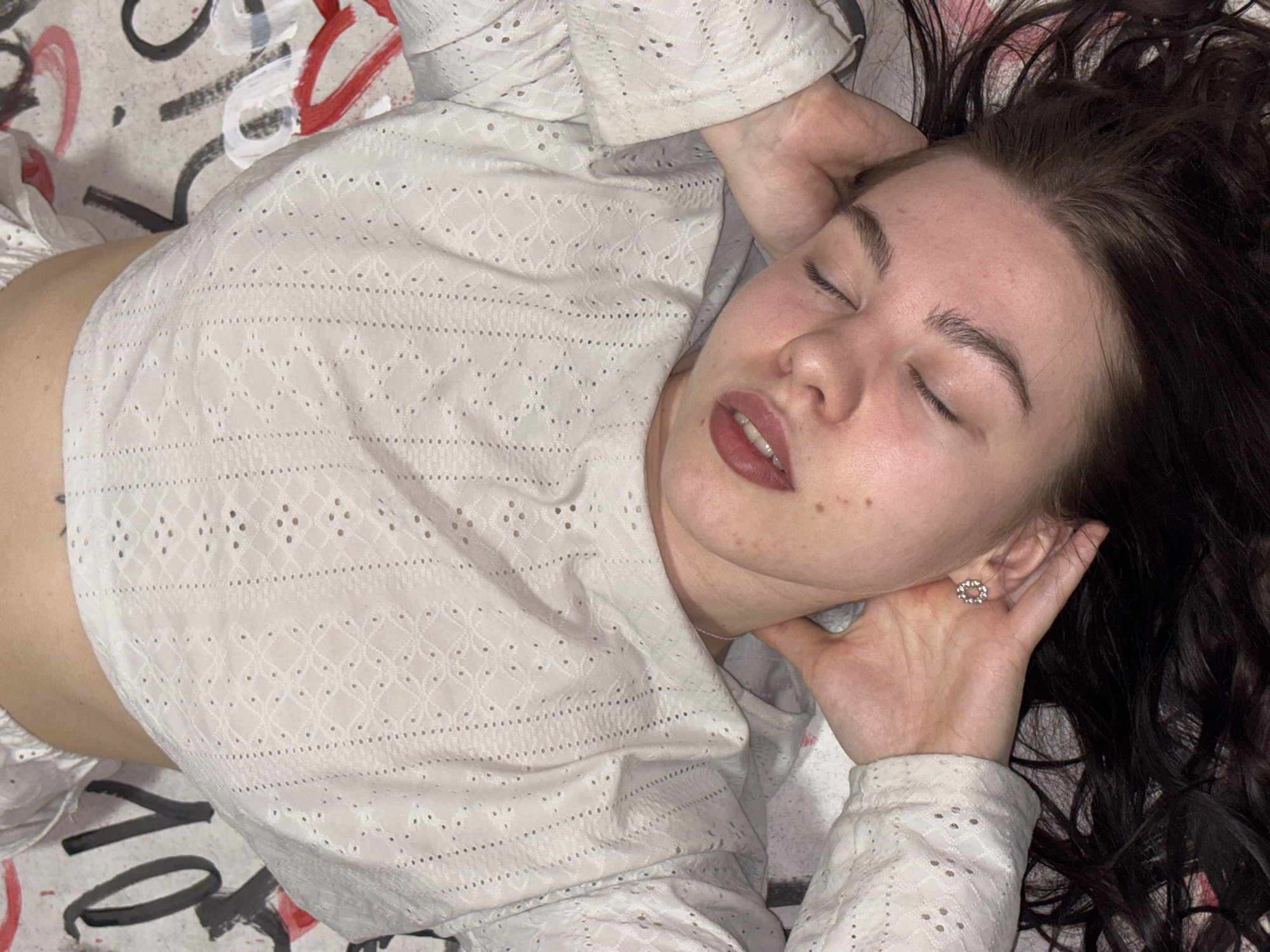 An overhead shot captures a young woman lying peacefully, eyes closed, on a surface adorned with abstract art. Her head is turned slightly to the viewer's left, with her dark, wavy hair fanned out around her. Her face shows a relaxed expression, with full lips slightly parted and natural-looking skin. She is wearing a white, long-sleeved top made of a delicate eyelet fabric with intricate geometric and dotted patterns, possibly a crop top, as a glimpse of her midriff is visible on the left.
Her right hand rests gently beneath her jawline, and a small, circular, silver-toned earring is visible in her right ear. The background surface is light-colored, possibly a bedsheet or blanket, featuring bold, expressive marks in black, white, and red, resembling graffiti or stylized brushstrokes. While some shapes hint at letters or numbers (like a large 'O' or 'C', and linear strokes), no discernible words or phrases are clearly visible. The overall atmosphere suggests a moment of rest or slumber indoors. The location context provided is Perm, Russia.