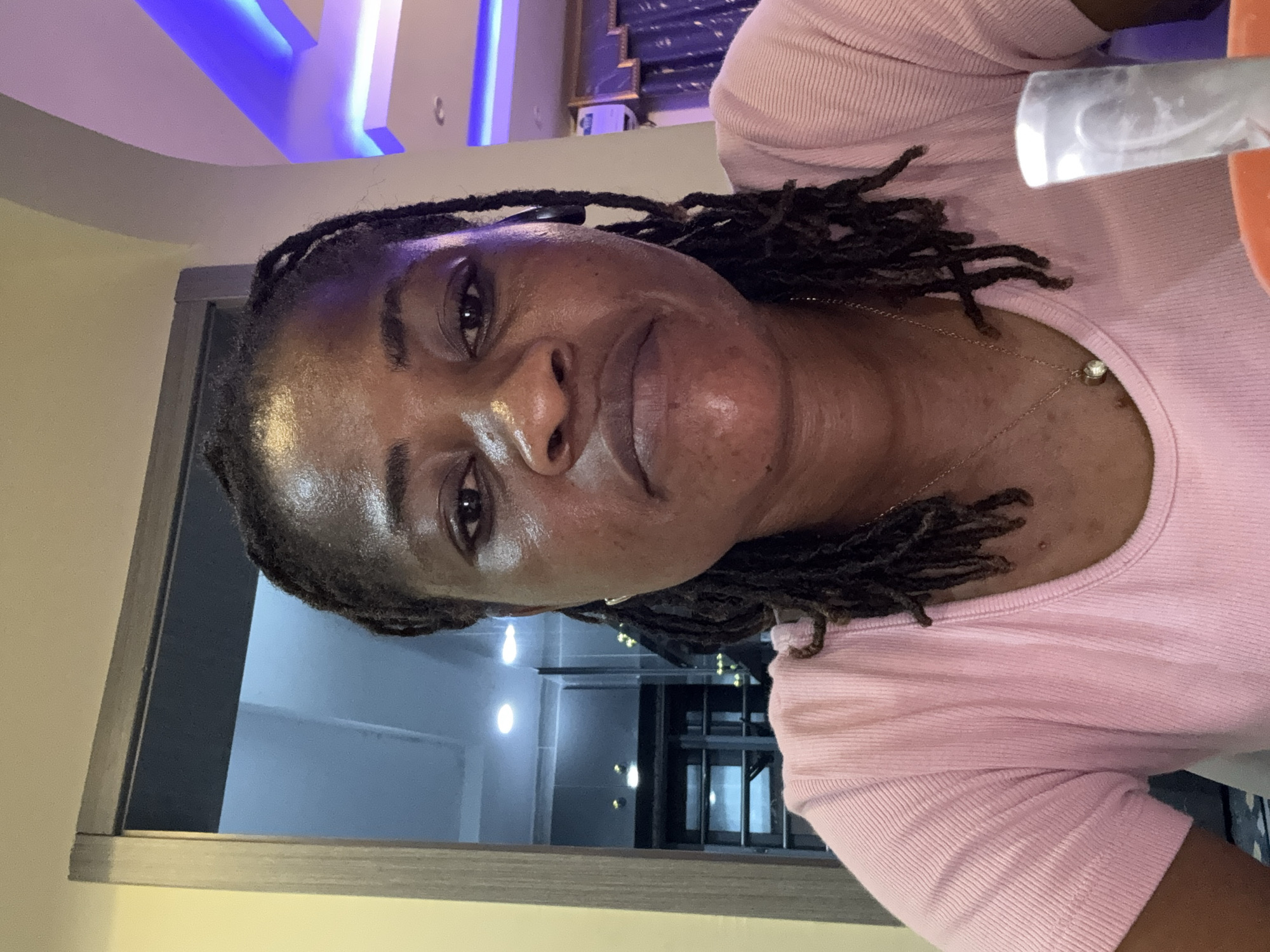 This is a medium shot, close-up selfie of a woman with dark skin and dreadlocks, looking towards the camera with a subtle smile. She is wearing a light pink, ribbed, short-sleeved top with a delicate gold chain around her neck that has a small, round, clear pendant. The lighting in the image appears to be artificial, possibly from overhead lights or lamps, as suggested by the glowing blue accents in the background and the general indoor ambiance. The background shows a modern interior with what looks like a doorway or partition in the foreground, leading to a more open space with fluorescent lighting. Reflections are visible in what might be glass doors or windows in the background. The overall impression is one of casual self-documentation, likely taken indoors in a contemporary setting in Ajara, Nigeria.