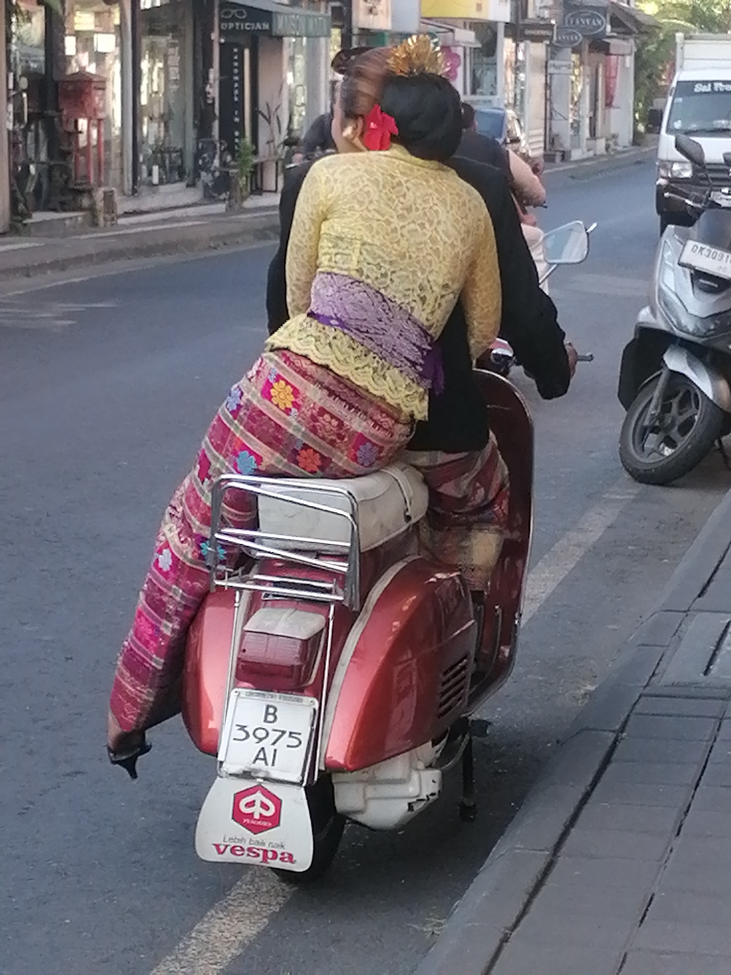 In Ubud District, Indonesia, a rear view captures a couple riding a red Vespa scooter. The woman in the back, adorned with traditional Balinese attire, wears a yellow lace top, a purple sash, and a vibrant pink and floral patterned sarong. She has a red flower in her hair and gold ornamentation. The man driving wears a dark top. The scooter displays a license plate reading "B 3975 AI" and a Vespa logo with the slogan "Lebih baik naik Vespa." The scene unfolds on a street with shops and buildings lining the background, and a paved walkway with a visible white line marking the road. Other scooters and a white van are also present in the vicinity.