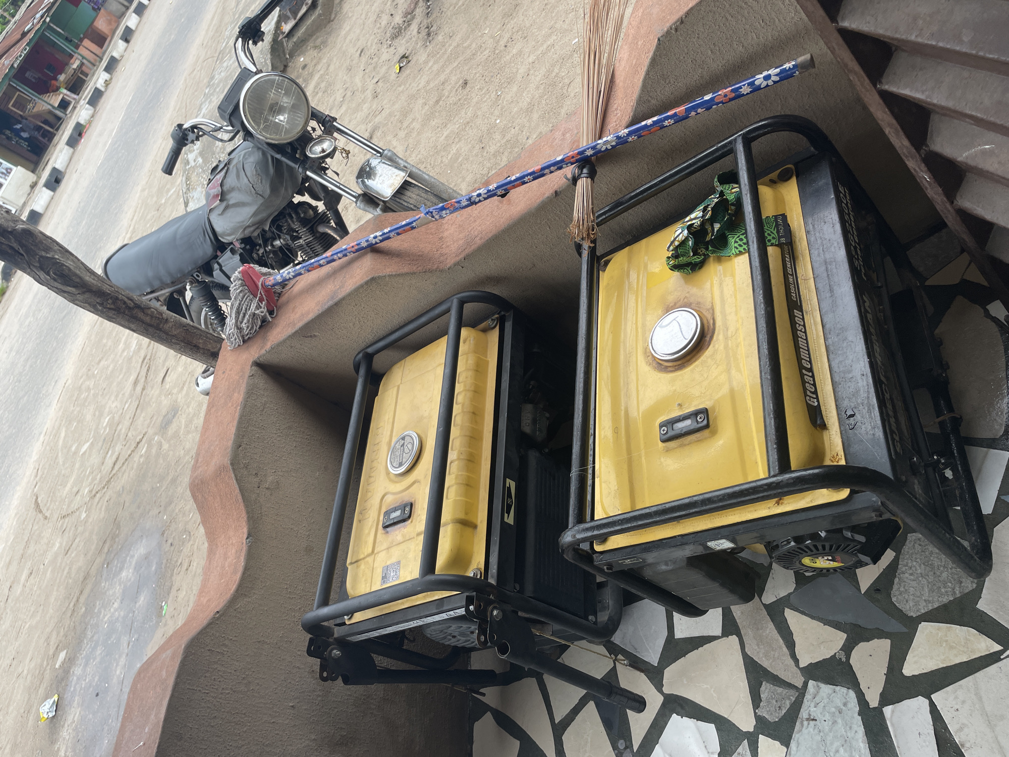 Two yellow generators with black frames rest on a marble-tiled surface. A blue floral-patterned mop pole and a broom lean against the generators. Above them is a motorcycle parked on a beige ledge. The motorcycle is grey and black. In the background is a street with buildings, characteristic of an urban setting in Badagry, Nigeria. The focus is on the generators and motorcycle, but the broader scene suggests a roadside or outdoor commercial context.