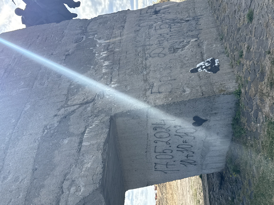 The image displays the lower section of a large, weathered grey concrete structure located in Izium, Ukraine. A strong, diagonal sunbeam cuts across the concrete from the upper right to the mid-left.
On the lower left face of the concrete, black spray-painted graffiti reads "17.05.2024" followed directly below by "10+10=10²." Beneath this text, a solid black heart symbol is spray-painted.
On the lower right face, additional black graffiti in Cyrillic script is present, with partially legible words including "Бро," "ДНИН БАЙДЯ," and "ТЕЛІНГГРОЙ." Below this text, a black and white stencil artwork depicts a crouched figure in a patterned robe.
The base of the concrete structure meets a paved surface of dark, irregular stones on the right, transitioning to dry earth with sparse green vegetation on the left. In the upper right background, a dark, indistinct sculptural element or figure is partially silhouetted against a partly cloudy sky.