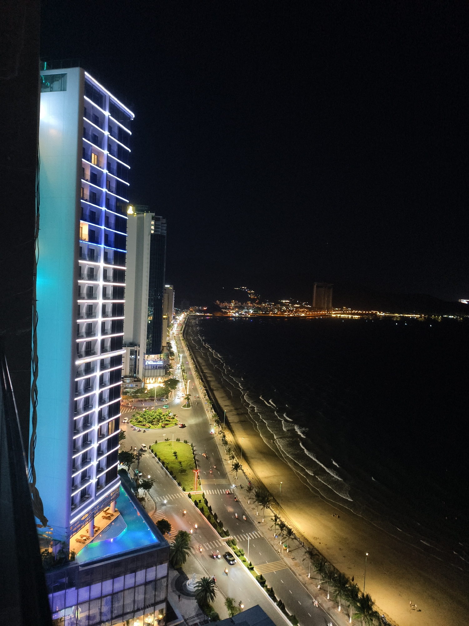 View from a high angle at night shows a beachside city. On the left is a tall hotel with blue and white lighting, next to a taller building. Below the hotel is a swimming pool. A street runs parallel to the beach. Lights are visible along the coastline. Further along the beach are more buildings and mountain-like terrain. The sky is dark. Cars and pedestrians are visible on the streets. The waves are visible along the shoreline.