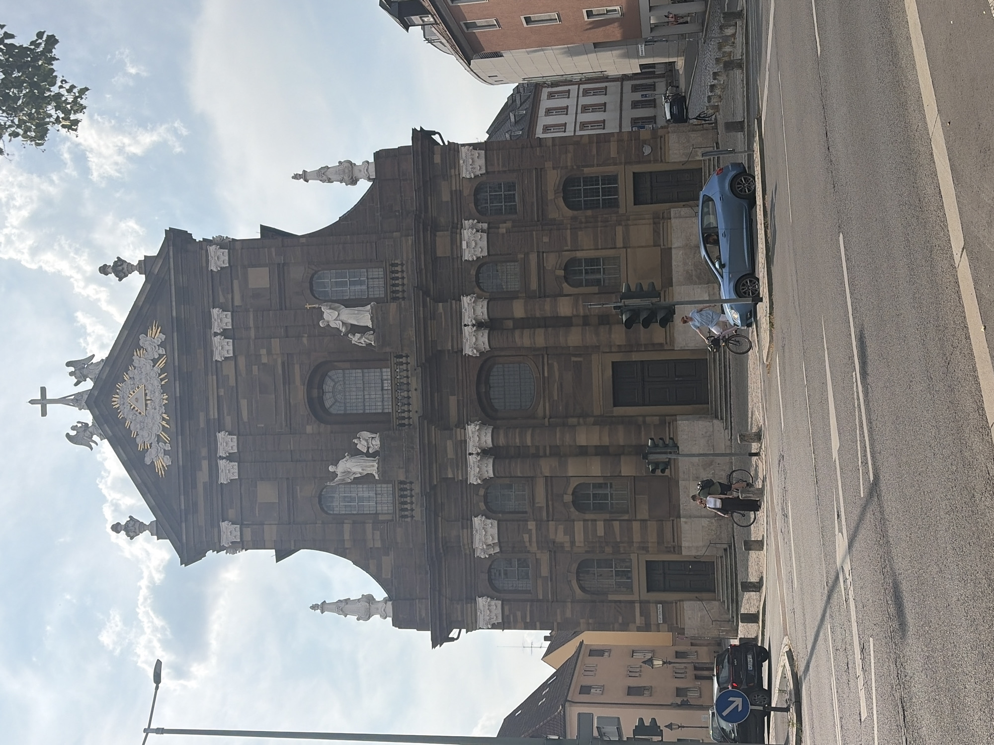 The image shows the exterior of the Würzburg Residence, a UNESCO World Heritage site in Würzburg, Germany. The grand baroque palace serves as the central backdrop. In the foreground, a street intersection is visible with asphalt road markings and pedestrian crossings. A light blue car is stopped at the intersection, with a person on a bicycle in the driver's seat, appearing to be waiting. To the left, another person on a bicycle is standing on the sidewalk near a traffic light. A black car is parked on the side of the road. Several traffic lights, bollards, and street lamps are present. The sky is partly cloudy. Buildings on either side of the palace are also visible, contributing to the urban setting.