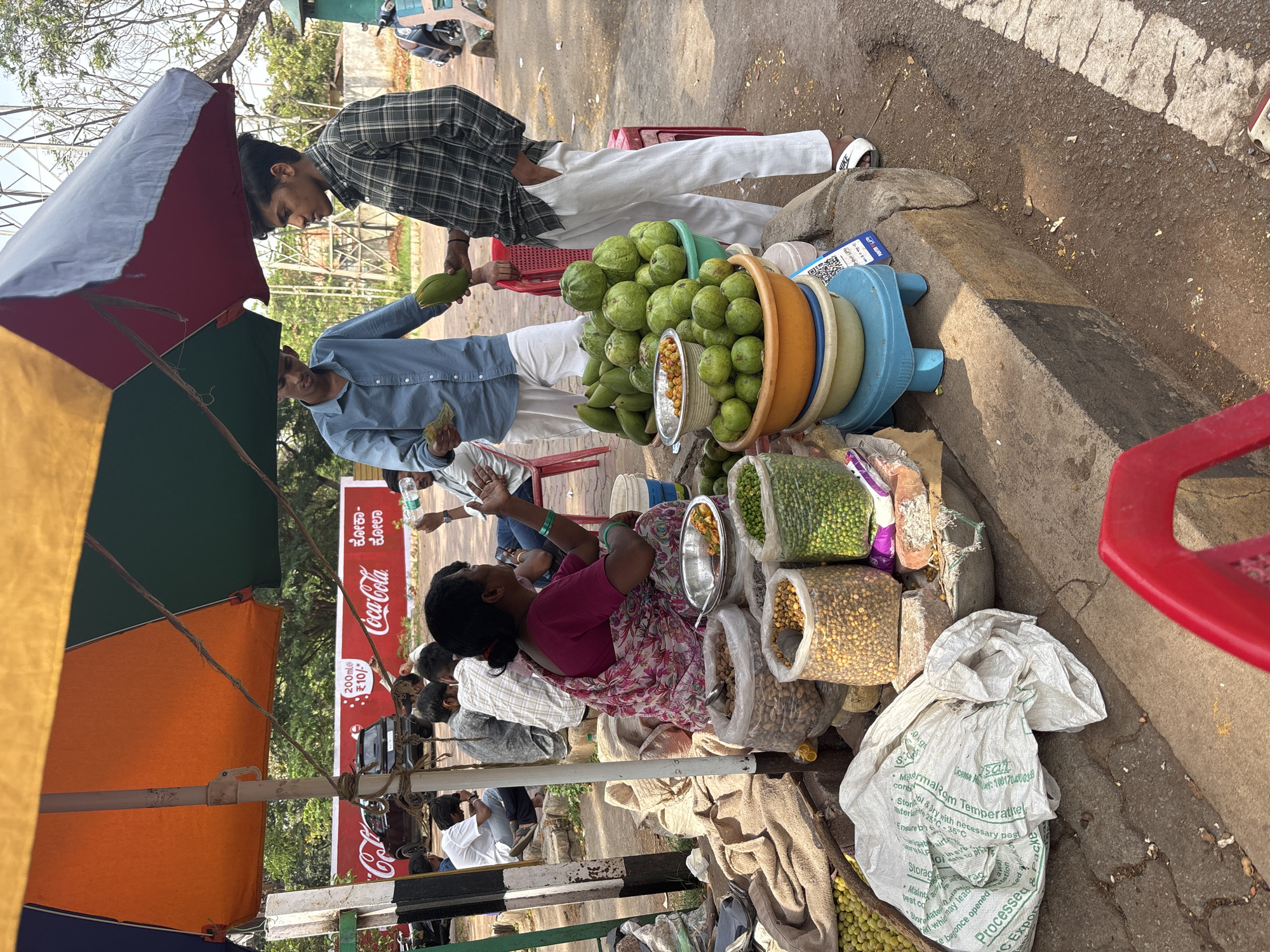 A street vendor in Hosapete, India, is selling fresh produce, including guava, peas, and nuts. Two men stand at the stall, one holding a guava and handing it to the other. The vendor, a woman, sits behind her stall, which is covered by a large umbrella. Coca-Cola advertising is visible on a sign near the vendor. People are sitting nearby. A motorcycle is visible in the background. The stall is situated on the roadside, next to a raised curb.