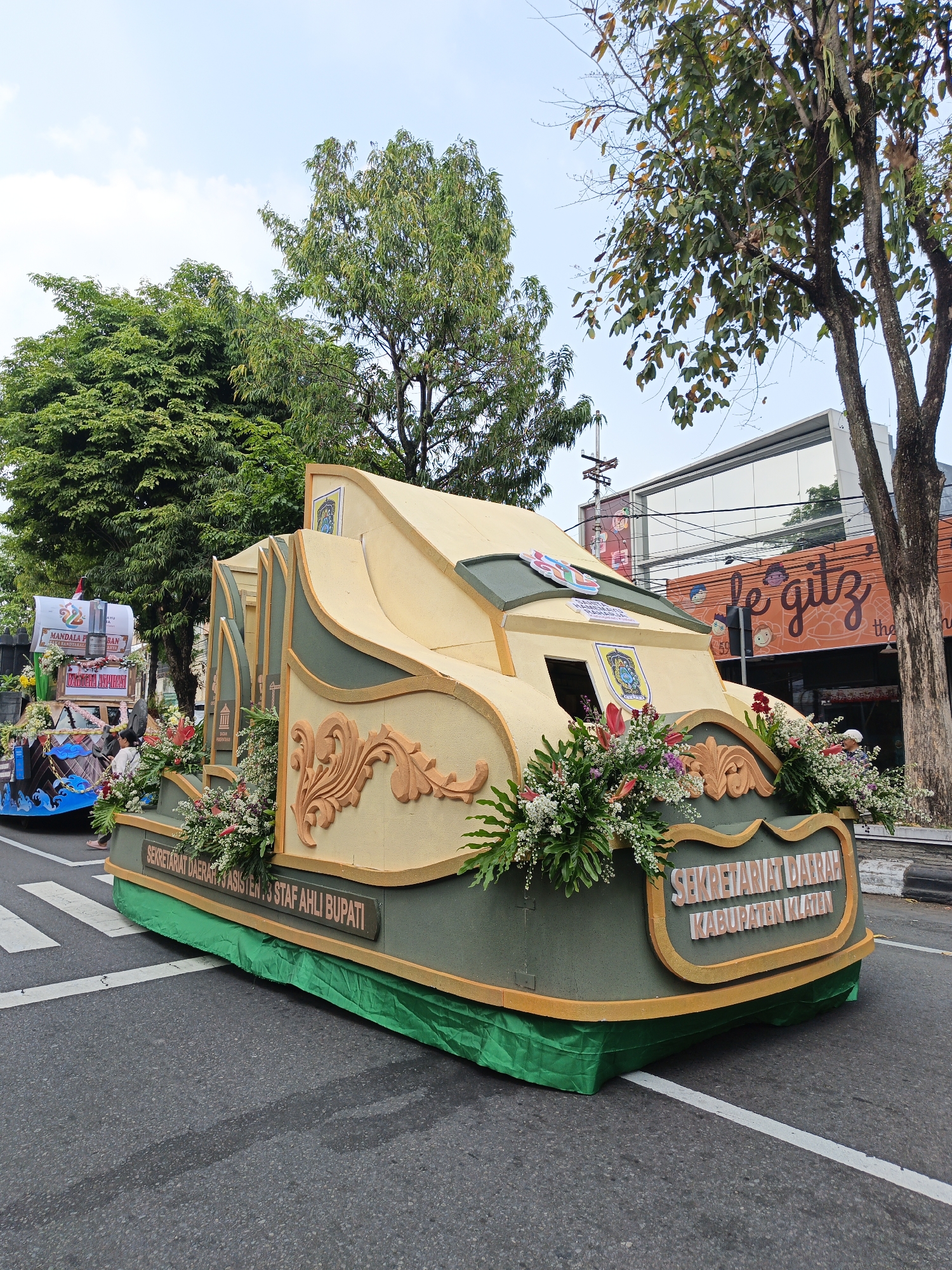 An outdoor, eye-level shot captures a parade float, designed to resemble a stylized building, traveling down a street in Klaten, Indonesia. The float is predominantly cream and dark green with ornate, gold trim. It is adorned with lush green foliage and colorful flowers, including red lilies and white and pink blossoms. Two signs are visible on the float: one in dark red letters reads "SEKRETARIAT DAERAH KABUPATEN KLATEN" and another, partially obscured, appears to read "SEKRETARIAT DAERAH ASSISTEN : 3 STAF AHLI BUPATI". A smaller emblem with a blue and gold crest is affixed to the side. In the background, buildings line the street, including a structure with an orange facade and the sign "le gitz" in large script. Trees with green leaves provide shade along the sidewalk. The street is paved and marked with white crosswalk lines. The sky is overcast with scattered clouds.