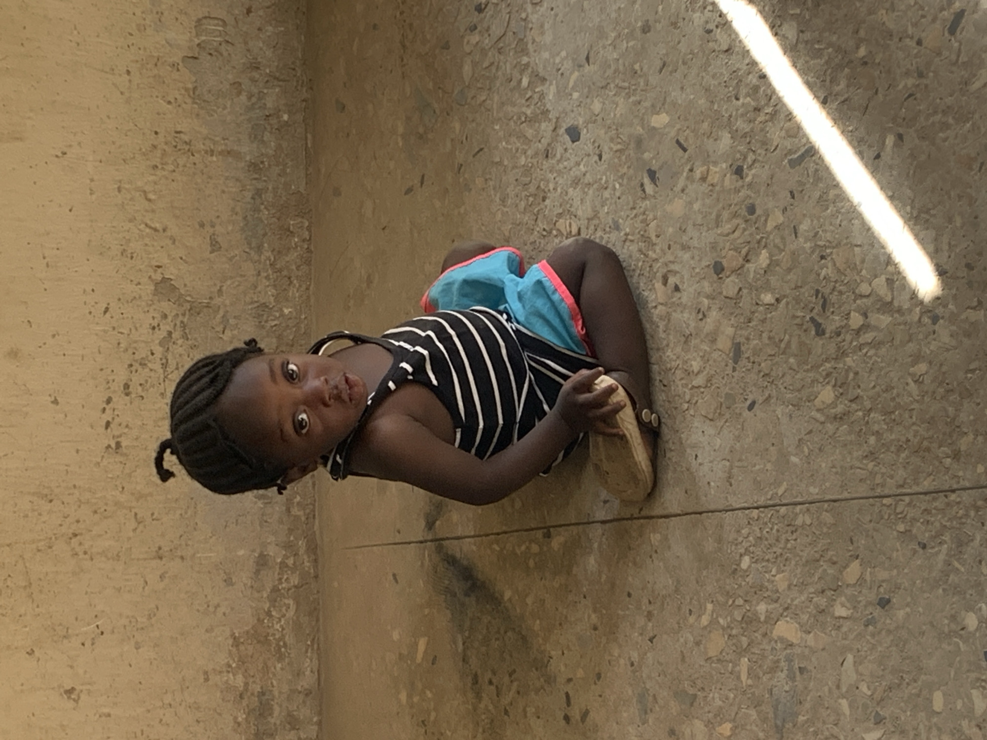 A young child is squatting on a speckled concrete floor in New Stadium, Nigeria. The child is dark-skinned, with braided hair pulled back into a small ponytail. They wear a striped tank top and blue shorts with a pink trim. One bare foot is visible, while the other is shod in a tan sandal. A light beam, possibly sunlight, illuminates the floor in the upper right corner of the image. A textured wall stands in the background. The child looks directly at the camera.