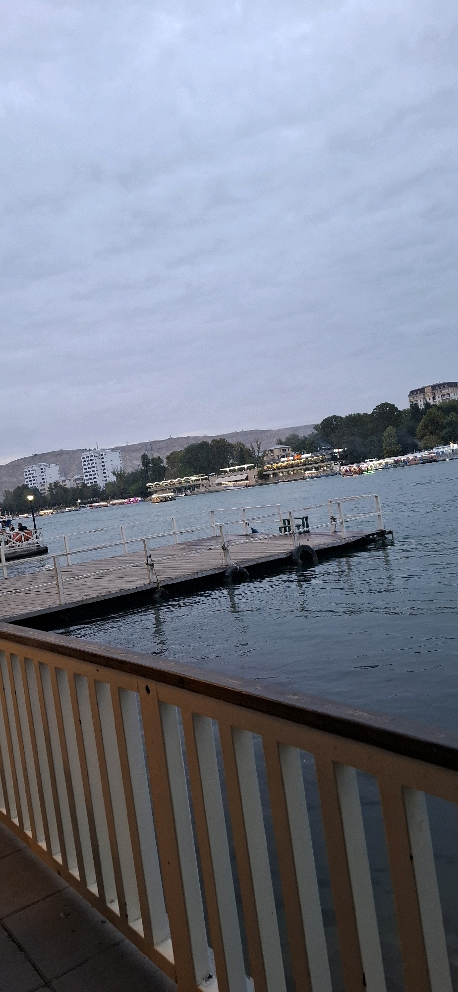 The image depicts a waterfront scene in the city of Mingachevir, Azerbaijan. In the foreground, a white and brown railing runs along the bottom of the frame, suggesting the viewer is on a walkway or balcony overlooking the water. A wooden pier extends into the dark blue water from the middle ground to the right. The pier has white railings and is equipped with black tires for boat docking. There are a few dark objects on the pier, possibly buoys or equipment. On the far left side of the pier, a small boat is docked. In the mid-ground, a body of water is visible, with several boats and what appear to be floating restaurants or entertainment venues lining the opposite shore. The architecture on the far shore includes several multi-story white buildings and smaller structures, all set against a backdrop of a gently sloping, barren hill. The sky is overcast and a muted gray, indicating a cloudy day or twilight. The overall atmosphere is calm and somewhat subdued.