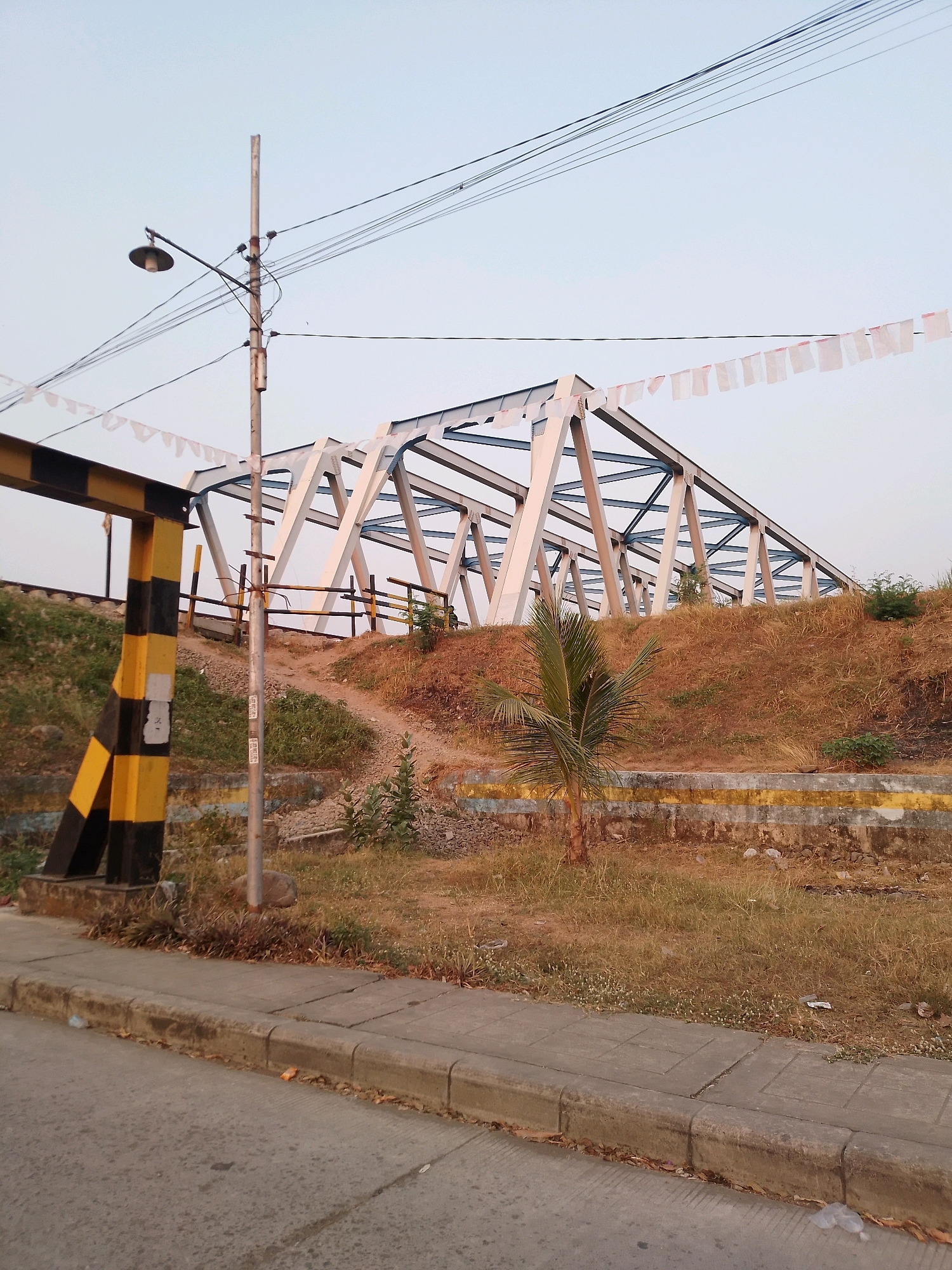The image captures a truss bridge in Kertosono, Indonesia, at dusk. The bridge, constructed with white and blue steel beams, spans over a grassy embankment. A black and yellow striped barrier is visible to the left, and a lamppost with wires stretches across the frame. Small white flags are strung along a wire above the bridge. The foreground shows a concrete sidewalk and a paved road. The embankment is covered in dry grass and some sparse vegetation. The sky is pale blue.