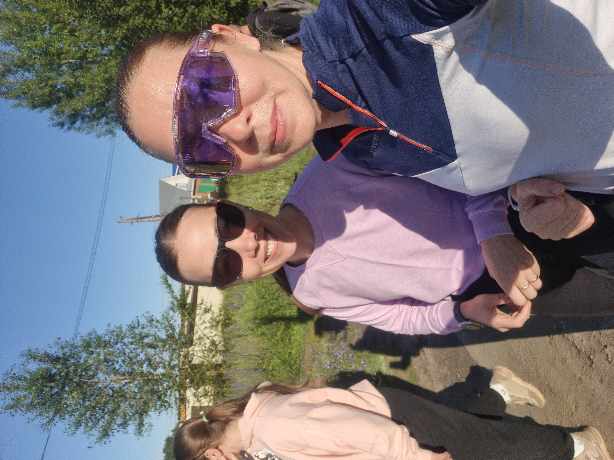 Two women are posing for a selfie outdoors in Chaikovsky, Russia, on a sunny day. The woman in the foreground is wearing large, transparent purple sports goggles and a navy blue and grey zip-up jacket with orange trim. She is smiling and looking at the camera. The woman behind her, to the left, is wearing black sunglasses and a light pink long-sleeved sweatshirt. She is also looking at the camera and smiling. A third person, a young woman with her hair in a ponytail, is partially visible on the far left, wearing a light pink hoodie and black pants. The background shows a dirt path, tall green grass and vegetation, trees, and a few buildings in the distance. The sky is clear and blue.