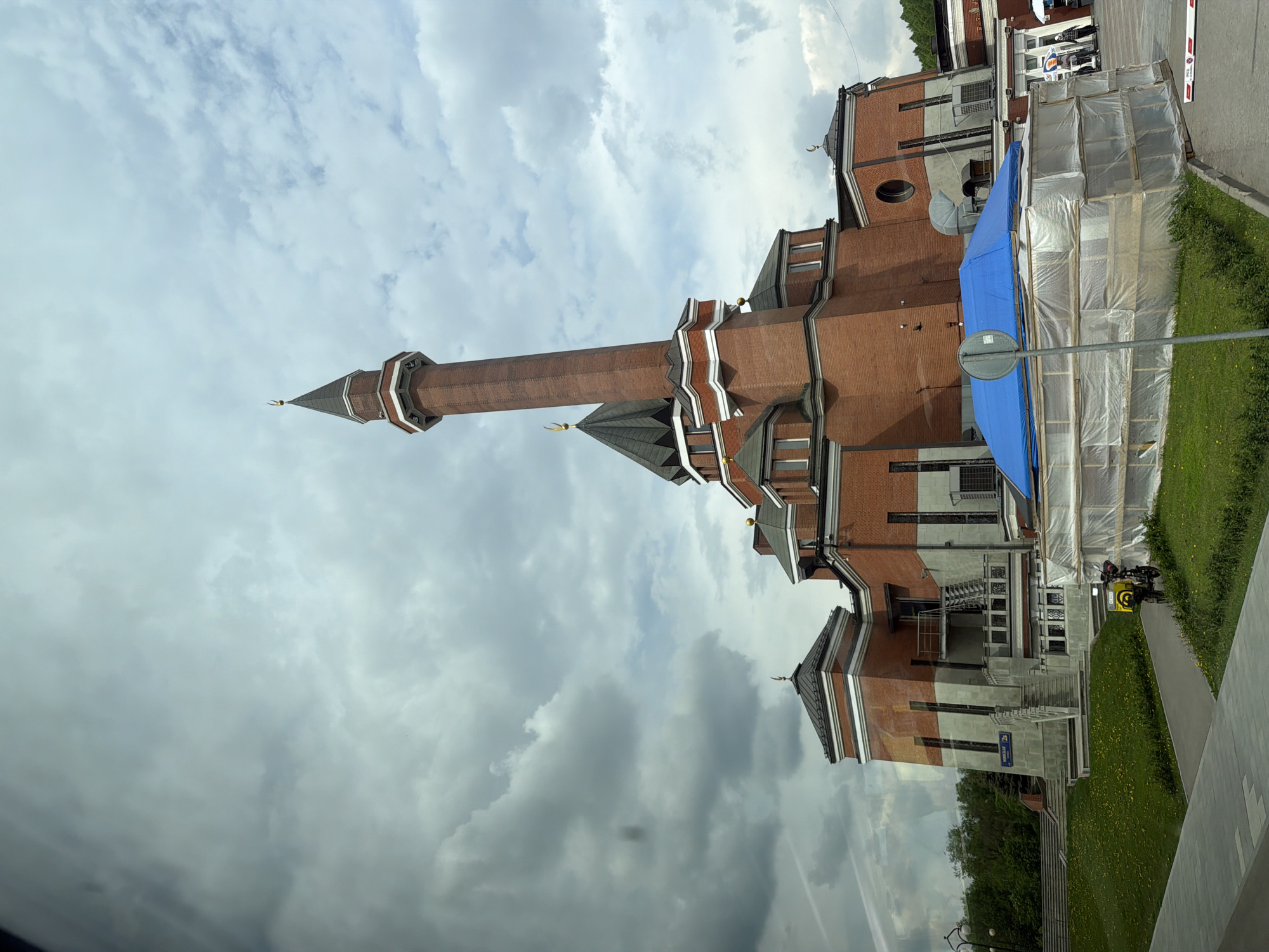 The image shows the Moscow Cathedral Mosque, a prominent mosque located in Moscow, Russia. The mosque features a tall minaret and red brick construction, indicative of its architectural style. Scaffolding and blue tarps are present, suggesting ongoing construction or renovation work. The surrounding environment includes green grass, trees, and a clear sky with cloudy patches, indicating an outdoor setting. There is a motorbike parked next to the building as well.