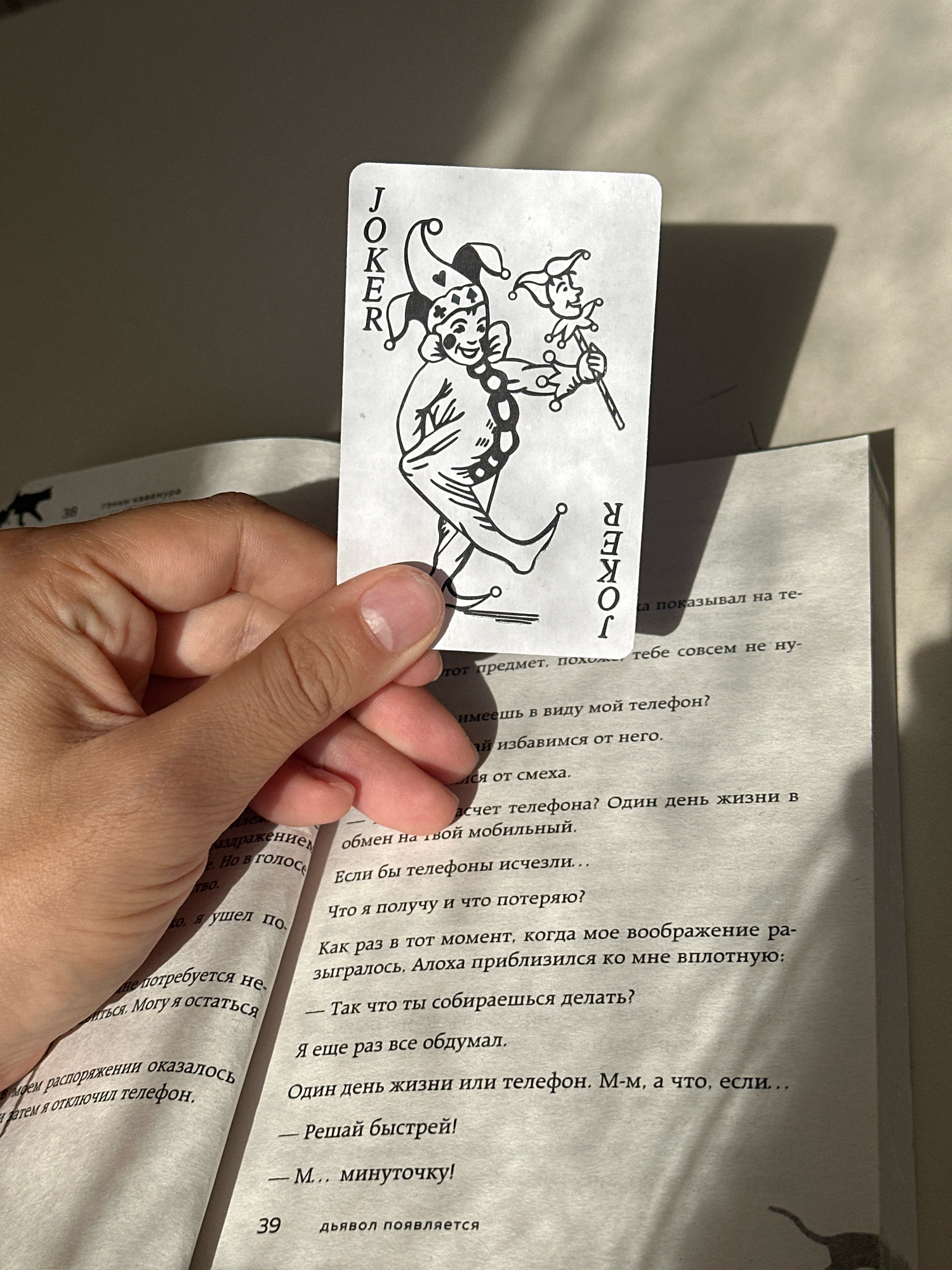 A hand holds up a "JOKER" playing card with a drawing of a jester against a book that is open. The card is held vertically, with the word "JOKER" written in a column on the left side. The book's pages are visible, with lines of text in Russian. The scene is lit from the side, casting a shadow of the card and hand on the wall behind. The image does not contain any information about the city of Gyumri, Armenia.