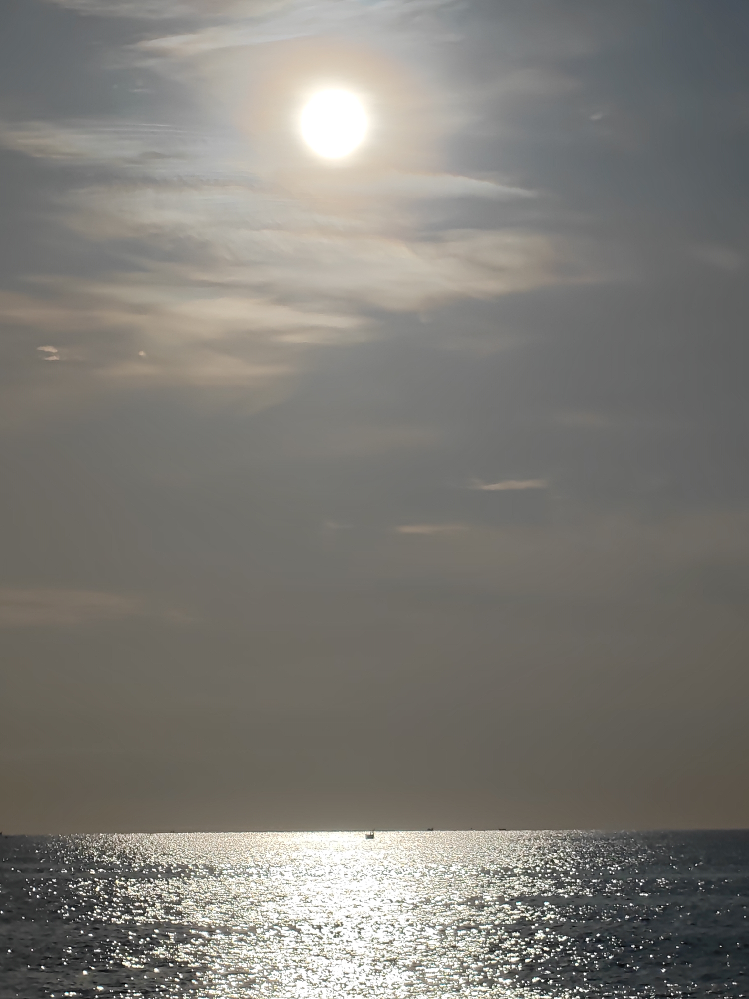 The image shows a seascape with the sun high in a hazy sky, likely taken in Lhokseumawe, Indonesia. The sun's glare dominates the upper portion of the frame, filtering through a layer of thin clouds. The majority of the image is filled with the expanse of the ocean, reflecting the sun's light with a shimmering effect. Several small boats are visible on the horizon line, appearing as dark silhouettes against the bright water. The scene is devoid of human presence except for the unseen occupants of the distant boats. The overall atmosphere is one of bright, hazy sunlight and vastness.
