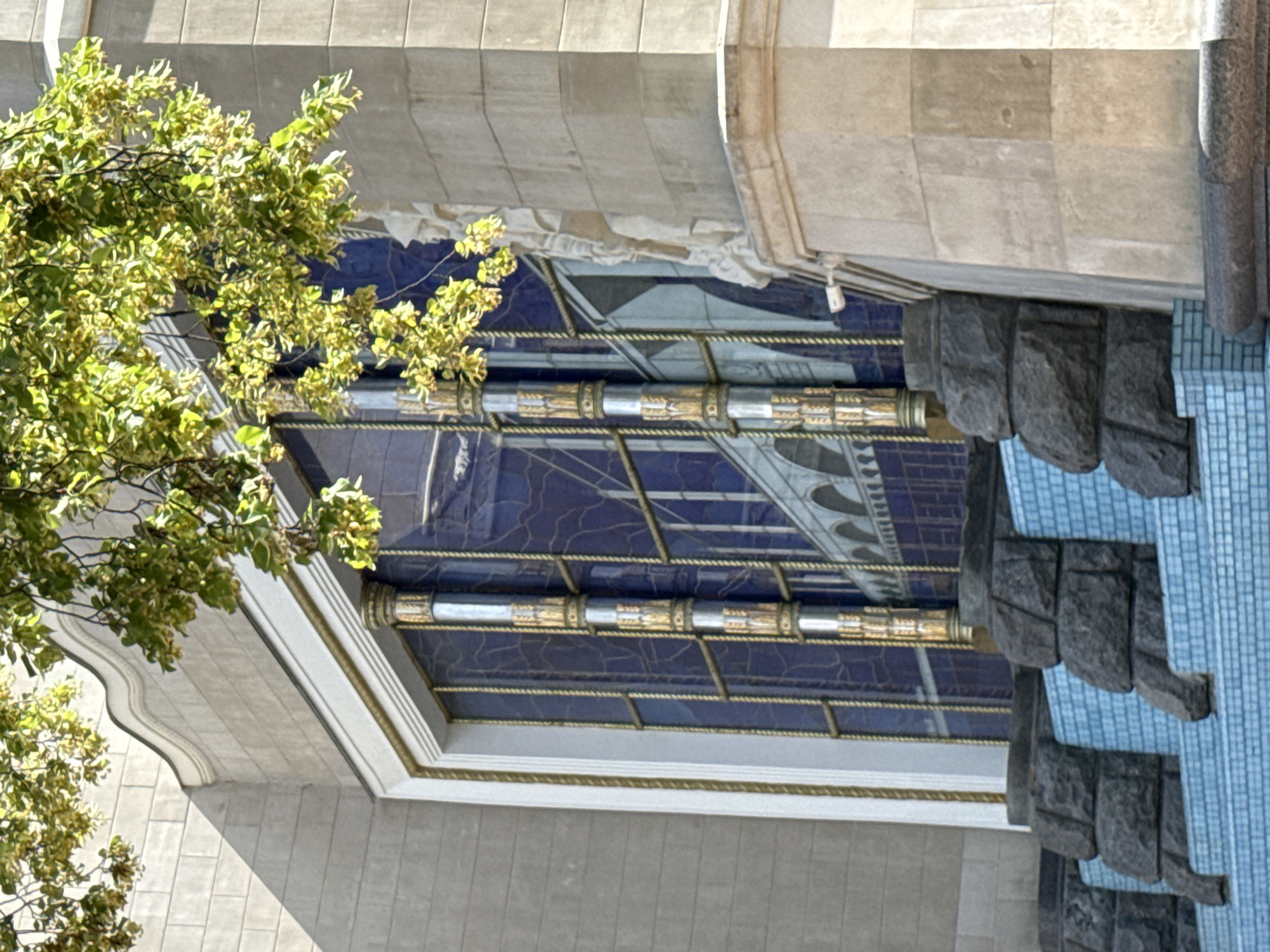 The image shows a close-up of a building with a large, decorative stained-glass window. The window features a deep blue background with a design of an arched bridge or aqueduct in white and gray. The window is framed by white molding and has vertical decorative elements that appear to be made of metal with gold accents. Below the window, there is a tiered structure made of dark gray stone blocks and light blue tiles. Lush green foliage from a tree hangs down from the top of the frame, partially obscuring the building. The building itself is constructed of light-colored stone blocks. The architectural style appears to be classical or art deco. The scene is bathed in sunlight, creating shadows on the building's facade. The specific location within Moscow, Russia, is not identifiable from the visual information provided. There are no people or actions depicted in the image.