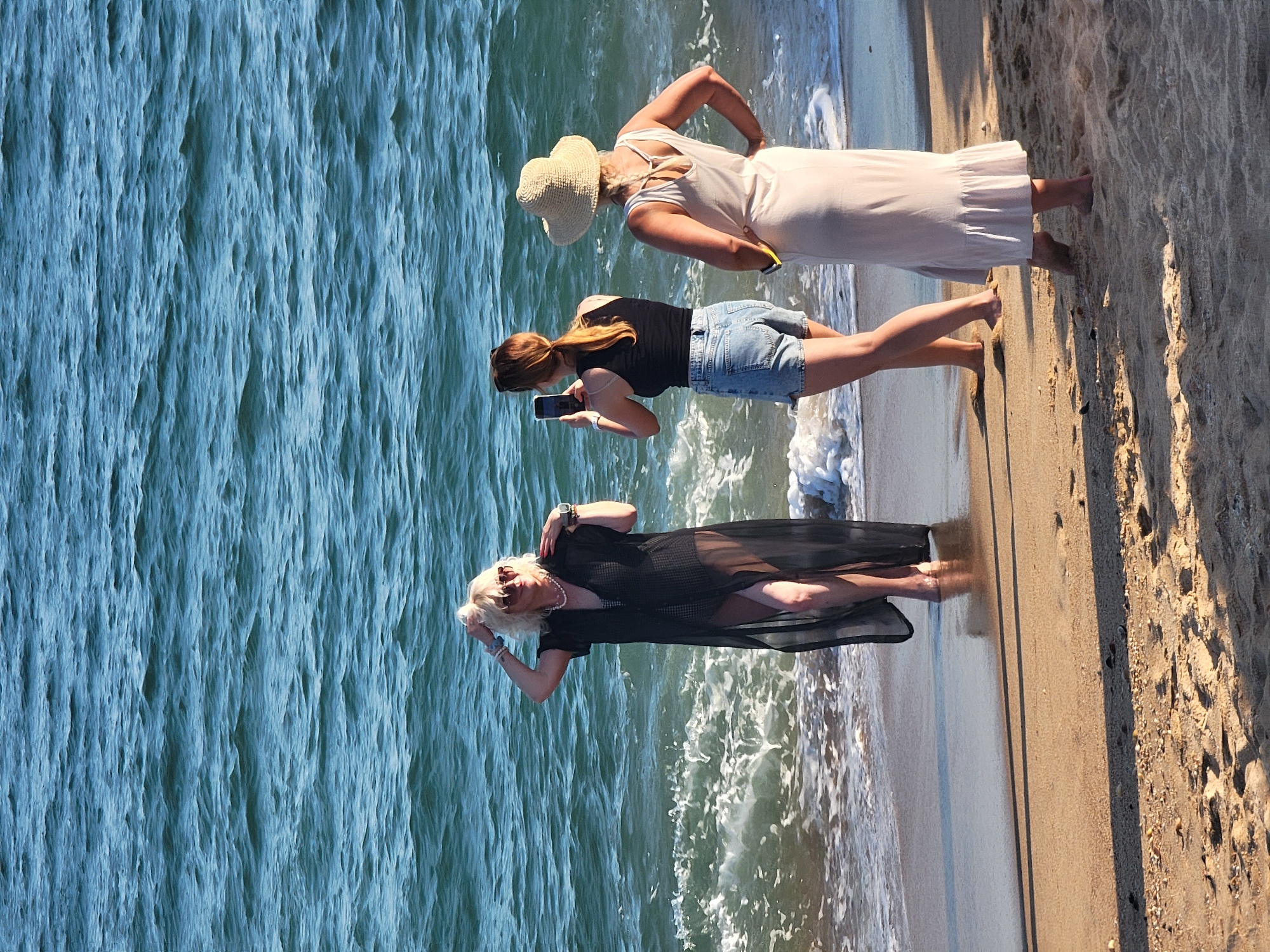Three women are on a sandy beach near the water's edge. The woman on the left, with blonde hair and sunglasses, is wearing a sheer black cover-up over a black swimsuit and is posing with her hand on her head. A second woman, with brown hair pulled back in a ponytail, is standing behind and to the right of the first woman, taking a photo of her with a mobile phone. She is wearing a black tank top and denim shorts. The third woman is standing to the right of the second woman, facing away from the camera. She has blonde hair and is wearing a light-colored, sleeveless dress and a straw hat. The ocean is in the background with gentle waves breaking on the shore. The sand is light brown with some footprints and small shells visible. The lighting suggests it is a sunny day. The scene takes place in Quarteira, Portugal, known for its beaches.