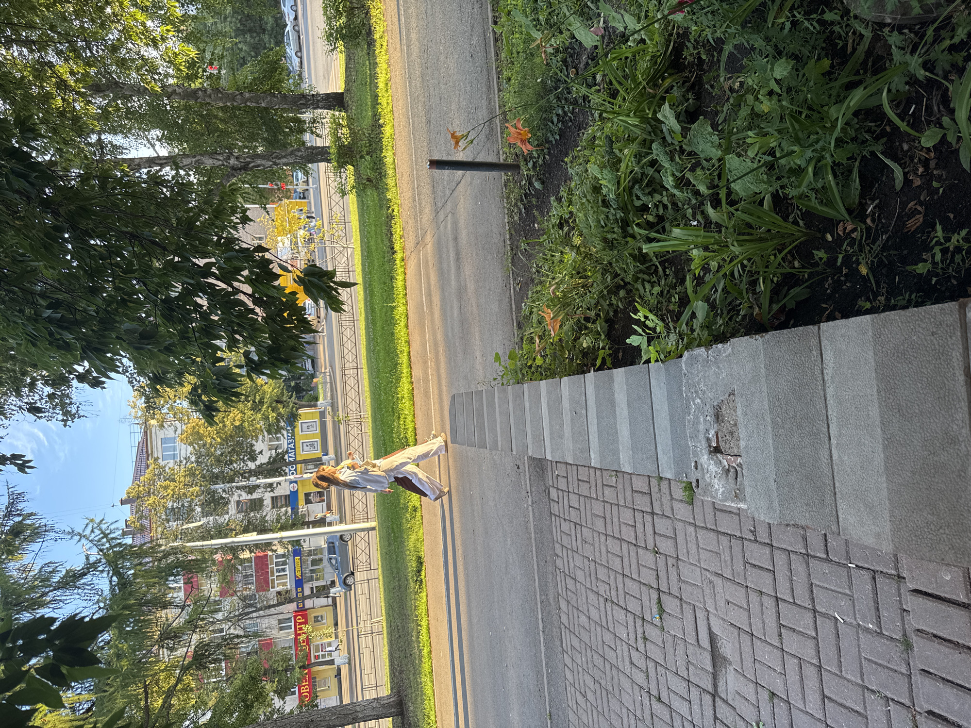The image shows a street scene in Ufa, Russia. The perspective is from above, looking down at a sidewalk and an adjacent grassy area. A woman is seen walking along the sidewalk, her back to the camera. She is wearing a light-colored shirt and pants. To the right of the sidewalk, there is a patch of greenery with some orange flowers, possibly daylilies. Further beyond the greenery, a road is visible with cars driving and parked. On the other side of the road, there are buildings, including a multi-story apartment building with yellow and white facades and red window frames. Some signs are visible on the buildings, including "РОСМАГАЗИН". The time of day appears to be late afternoon or early evening, with the sun casting long shadows and illuminating the scene with a warm, golden light. The sky is clear and blue. The overall impression is a quiet, everyday moment in a city environment.