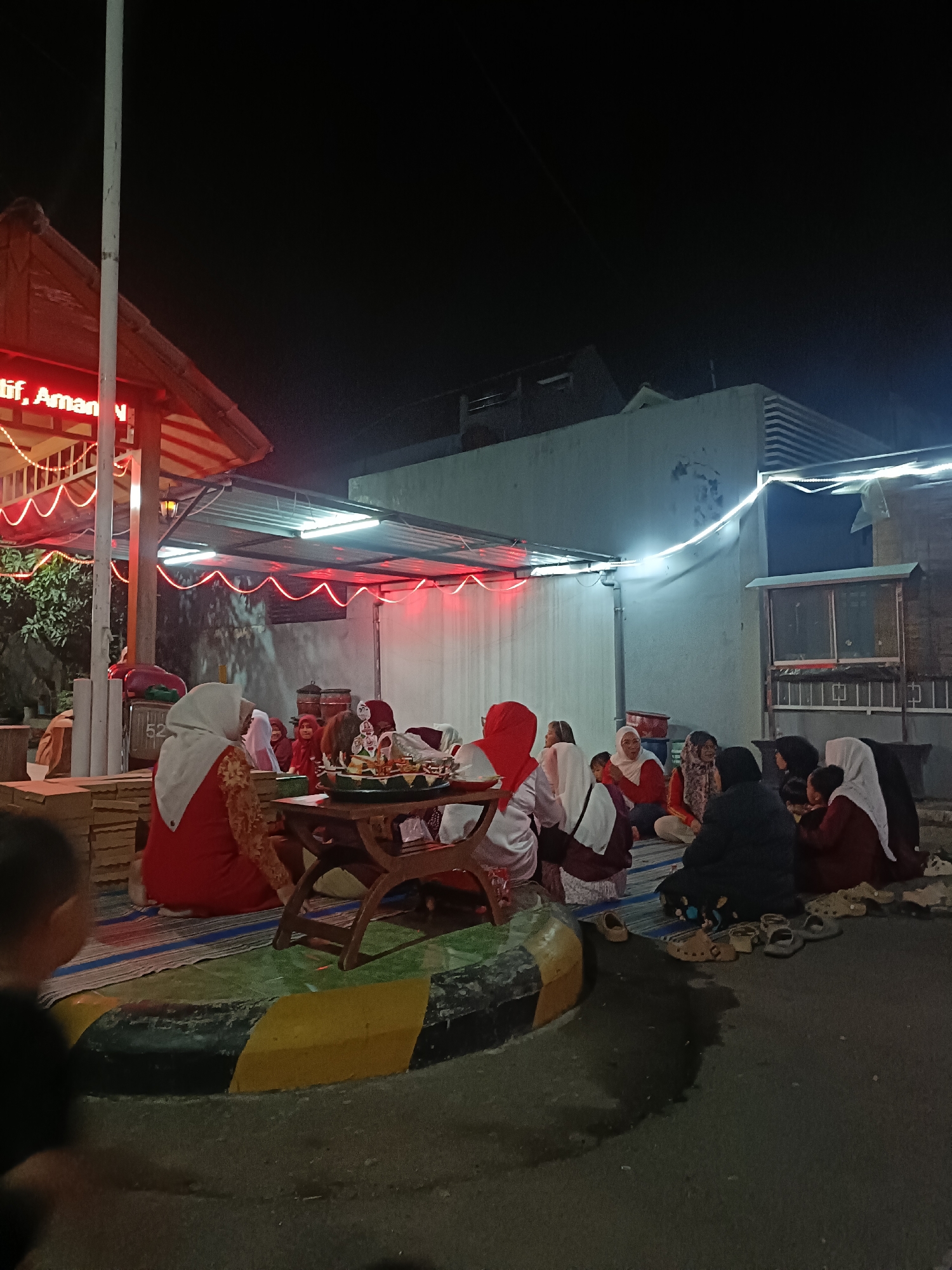 A group of people, predominantly women and children, are gathered on mats on the ground at night, likely outdoors in an urban setting, possibly in Kediri City, Indonesia. They appear to be engaged in a communal activity around a low wooden table with a tiered food display. Many of the individuals are wearing Islamic attire, including hijabs and long dresses, suggesting a cultural or religious event. Red and white string lights are visible, illuminating the area. In the background, a building with a sloped roof and outdoor lighting is present, along with a white wall and what appears to be a fence or gate. The ground is paved, with a distinct yellow and black striped curb. Several pairs of sandals are scattered on the ground, indicating they have been removed. The overall atmosphere suggests a gathering for a meal or celebration.