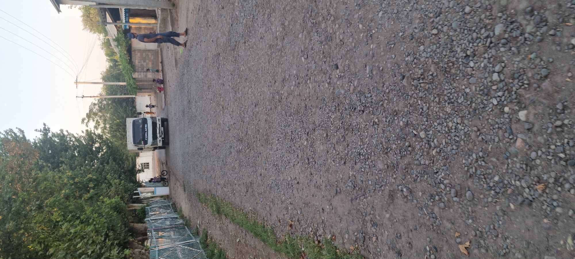 The image depicts a street scene in Хаваскор, Tajikistan, characterized by a gravel road leading into the distance. On the left, a decorative metal fence borders a grassy area with trees. Further down the road, a white truck with the word "FORLAND" on its windshield is parked. Several children are visible, some playing near the truck, and one is on a bicycle. On the right side of the road, a man in a dark blue t-shirt and jeans walks away from the camera. Behind him, a building with a flat roof and cinder block walls is visible, and further down, another building with a more traditional architectural style. Power lines are strung across the sky. The overall environment appears to be a rural or semi-urban residential area.