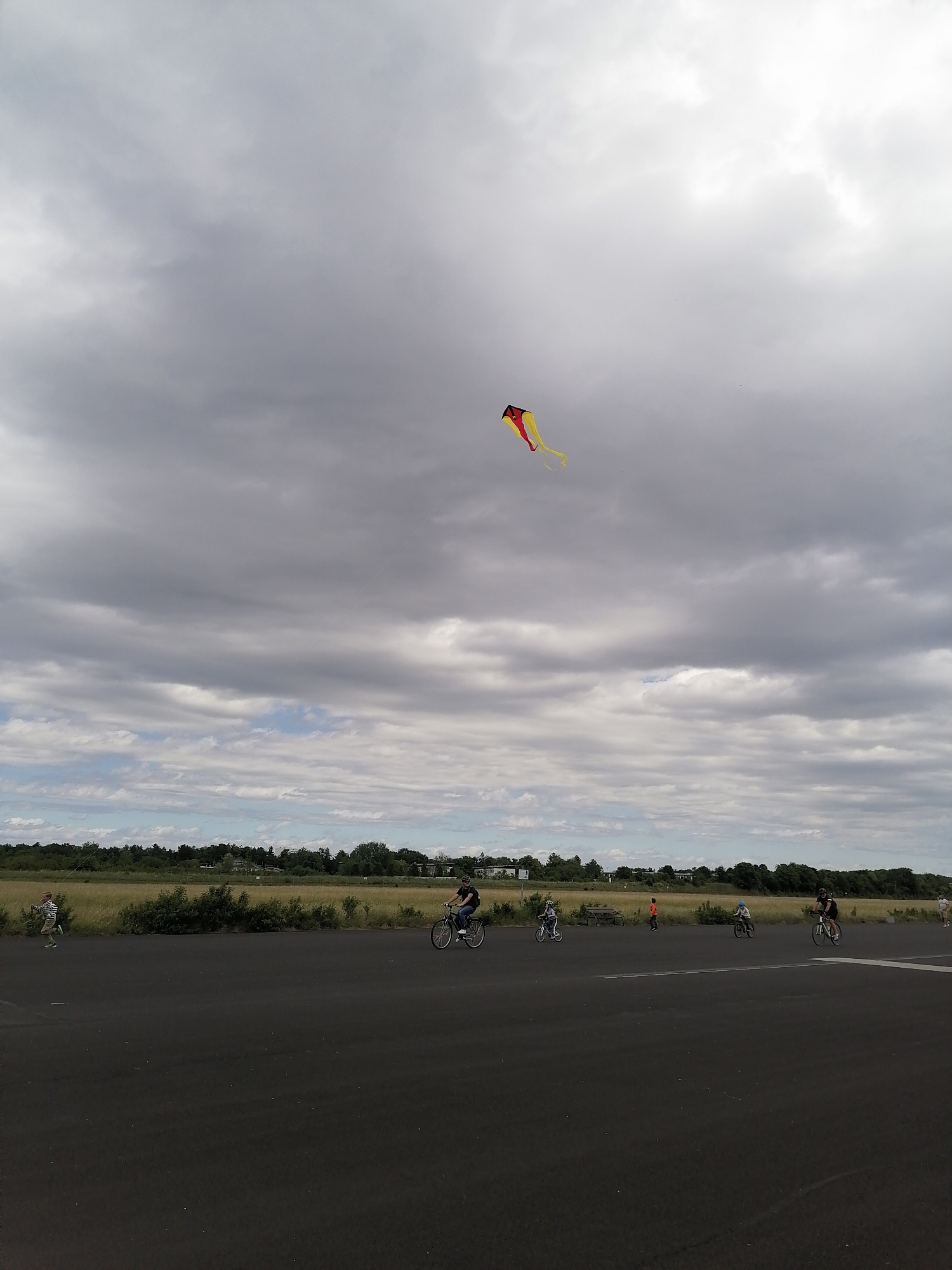 This is a wide-angle, medium shot of a park in Unterhaching, Germany, on a cloudy day. In the distance, a red and yellow kite is flying in the overcast sky. In the foreground, a man on a bicycle is riding on a paved surface. To the left of the cyclist, a child is running, holding a kite string. Further in the distance, other people are visible on bicycles and running, suggesting a recreational activity. The background features a field of dry grass, trees, and some buildings.