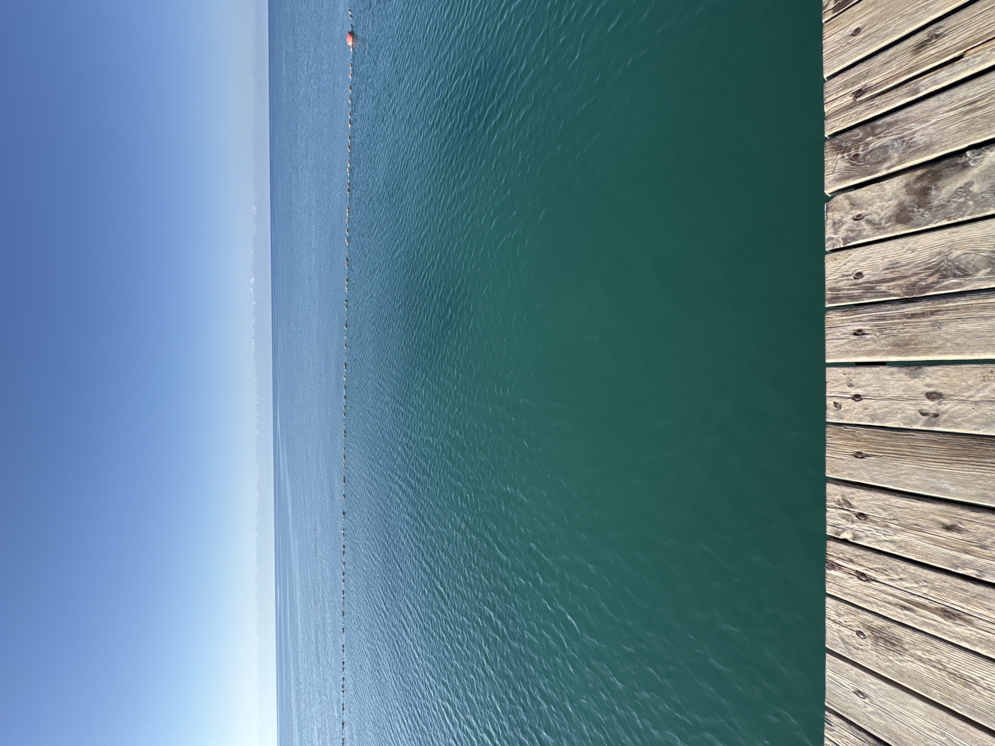 A wide, clear blue sky is above a calm, turquoise body of water, possibly a lake or sea, with a distant hazy shoreline. A thin line of small buoys stretches horizontally across the water. In the foreground, a wooden dock made of aged planks runs along the bottom of the frame. The scene is serene and devoid of people or any activity. The location is identified as Sary-Oy, Kyrgyzstan.