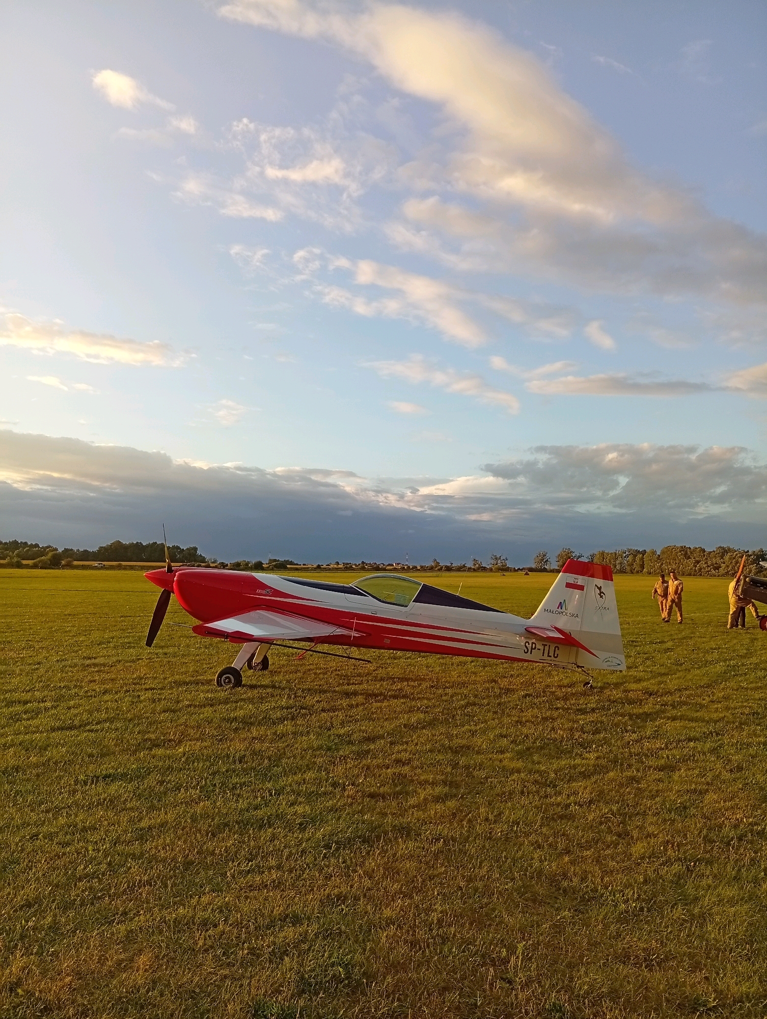 A red and white Extra 330SC aerobatic aircraft is parked on a grassy airfield in Szewce, Poland. The aircraft, with the registration SP-TLC visible on its tail, is angled to the right, with its propeller facing the viewer. The sky above is partly cloudy with patches of sunlight. In the background, a line of trees forms the horizon, and several figures in tan jumpsuits stand on the grass, appearing to be in conversation or attending to another aircraft. The lighting suggests it is late afternoon or early evening.
