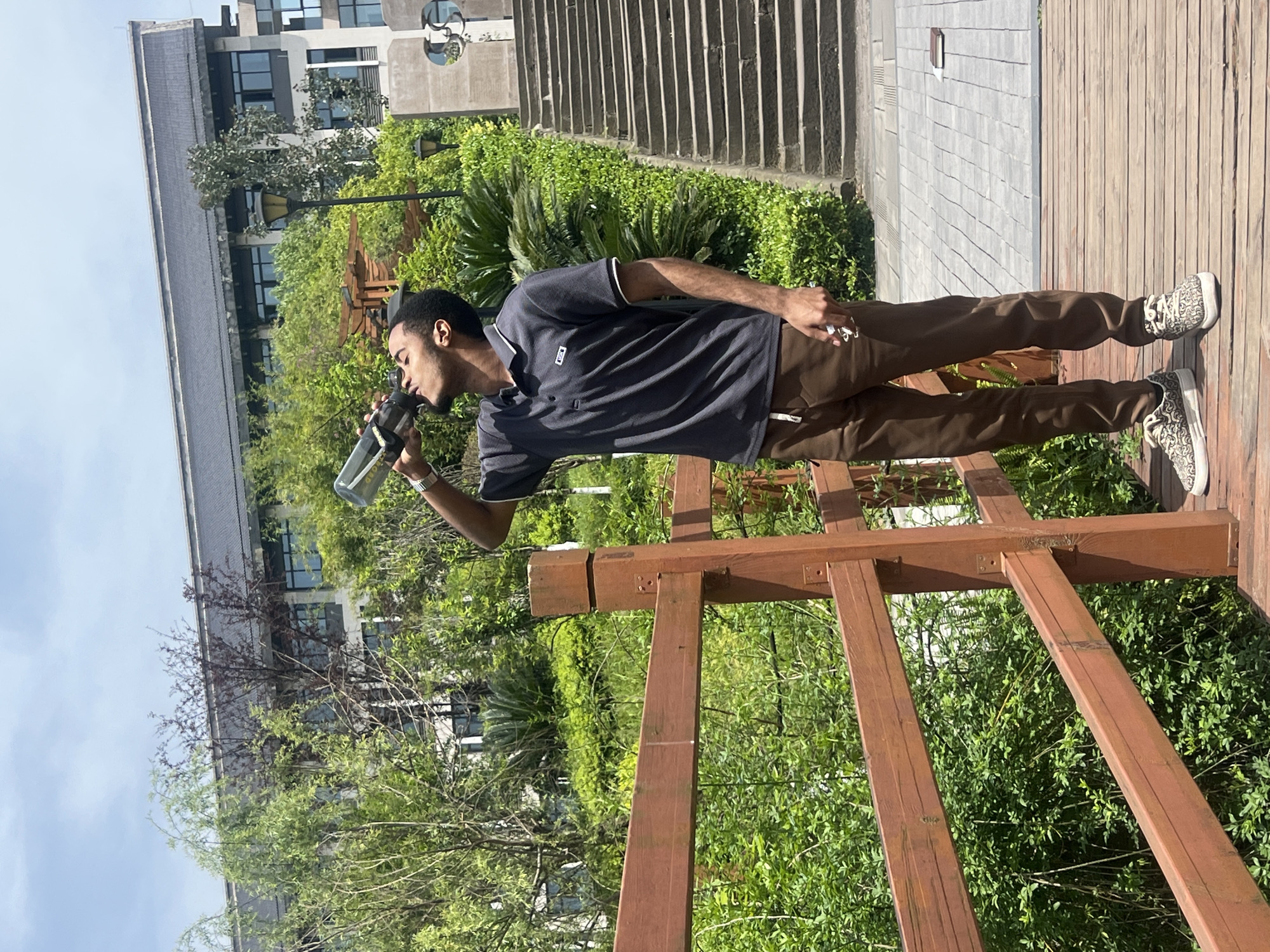 A man stands on a wooden platform in Jiangjin, China, drinking from a clear water bottle. He is wearing a short-sleeved polo shirt and brown pants, with patterned sneakers. A wooden structure, possibly a trellis or pergola, is near him, and there is lush green foliage in the background. Buildings are visible in the distance, suggesting an urban park or garden setting within the city. Stairs are also visible, leading to an upper level.