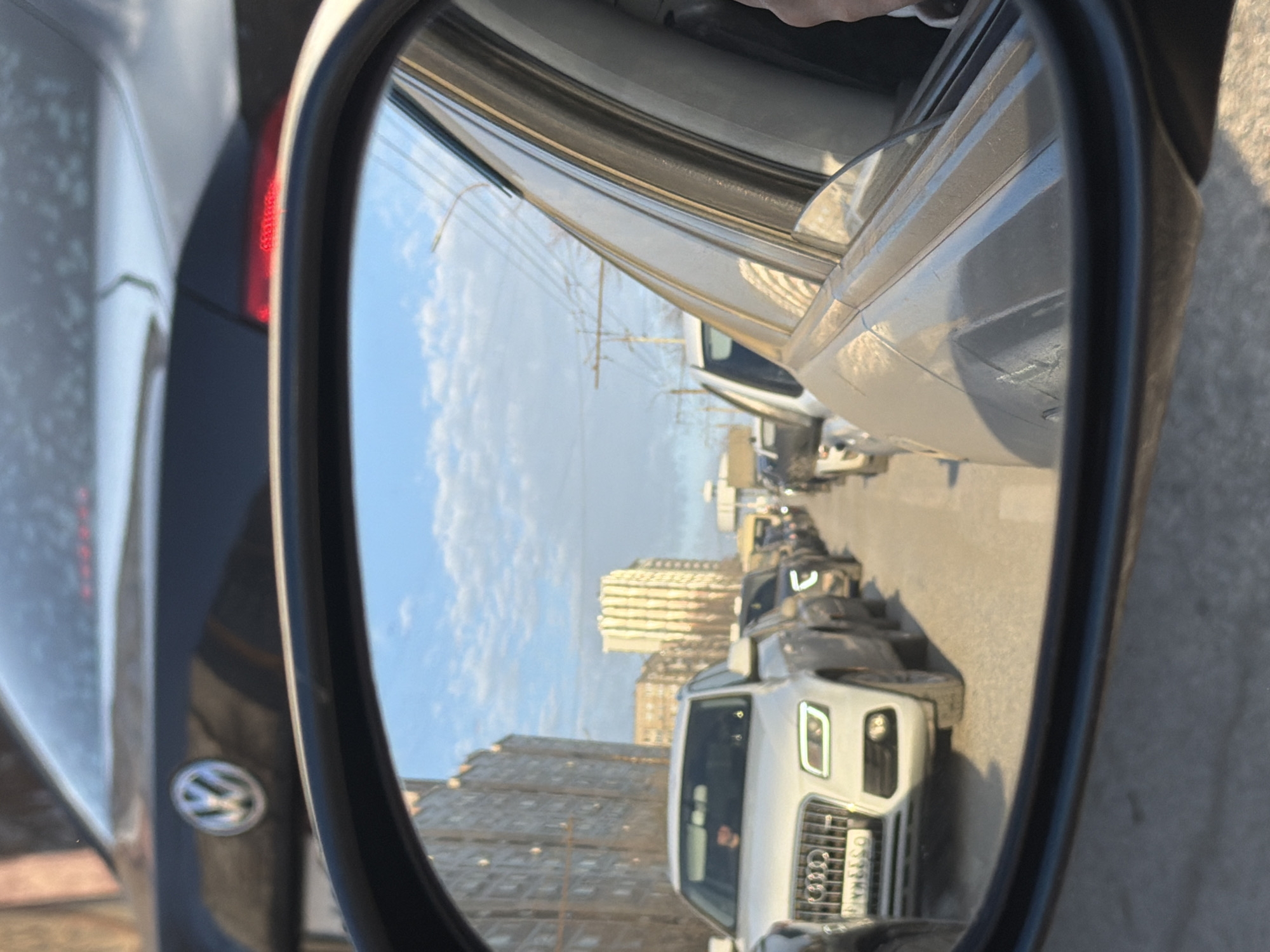 The image is a view from a car's side mirror showing a traffic jam in Izhevsk, Russia. Reflected in the mirror are several cars, including a white Audi Q7 with license plate “O553TK18”. Buildings line the road in the background, and the sky is partially cloudy. The car taking the picture appears to be a Volkswagen. The overall environment is urban and congested.