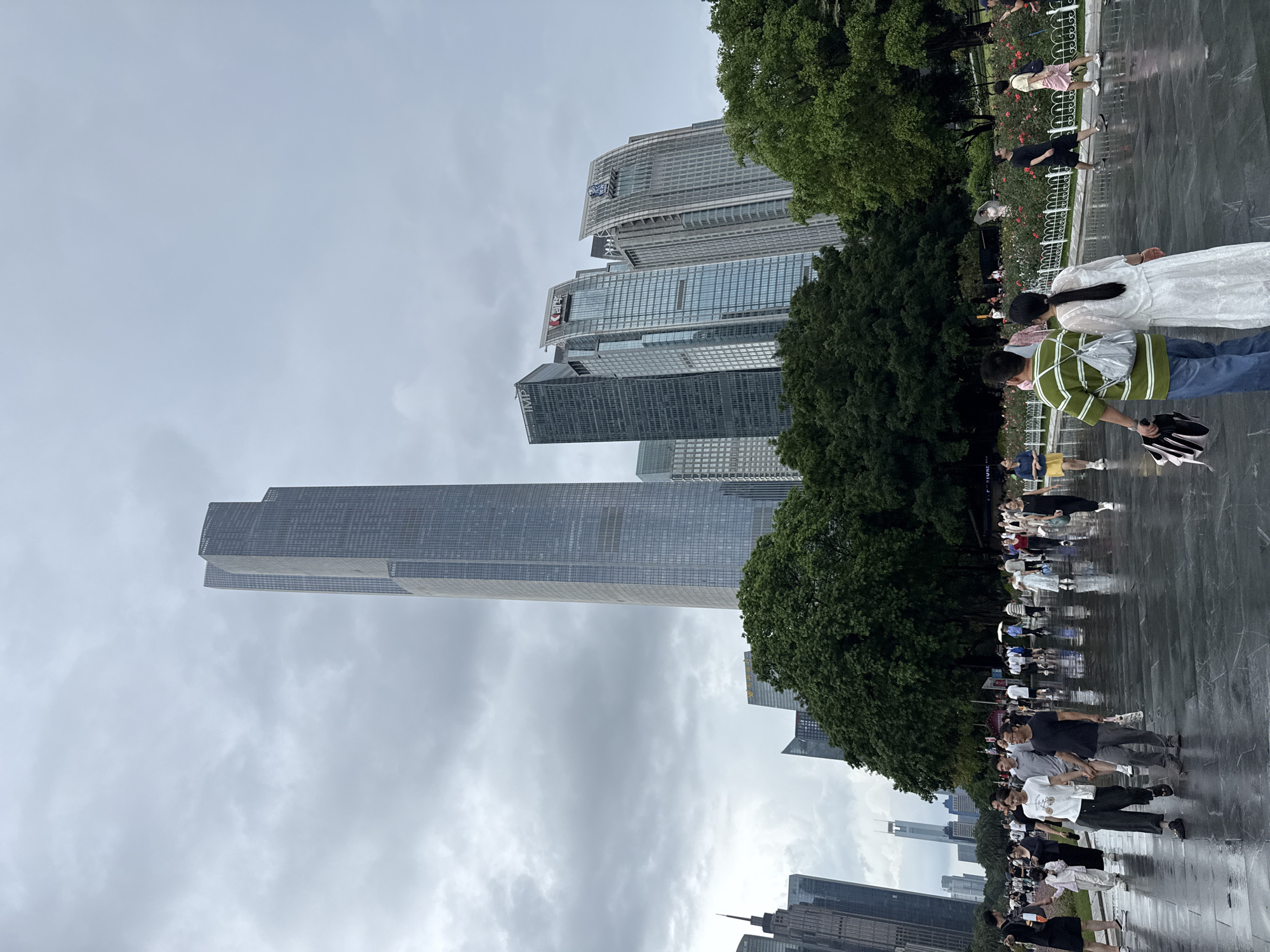 A wide-angle, eye-level shot depicts a cloudy day in Tianhe District, China, with a paved, wet walkway in the foreground. Numerous people are walking, some in pairs, and others individually. The walkway leads to a park with lush green trees and a rose garden. In the background, several modern skyscrapers dominate the skyline, with the tallest building featuring a distinctive stepped design. The sky is overcast, with dense grey clouds.
