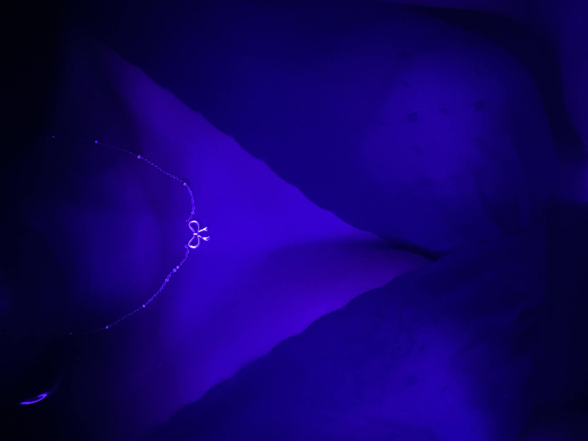 A close-up shot displays a delicate silver necklace with a bow-shaped pendant against a dark blue textured surface. The necklace extends diagonally across the frame from the top right, where the bow pendant is positioned, towards the bottom left. The surface appears uneven, possibly fabric or skin, with variations in tone and shadows suggesting depth. The lighting is predominantly blue, obscuring detail and giving the scene a cool tone. There are no people visible in the image. The image is not necessarily located in Maigizo, Nigeria; no information supports this.