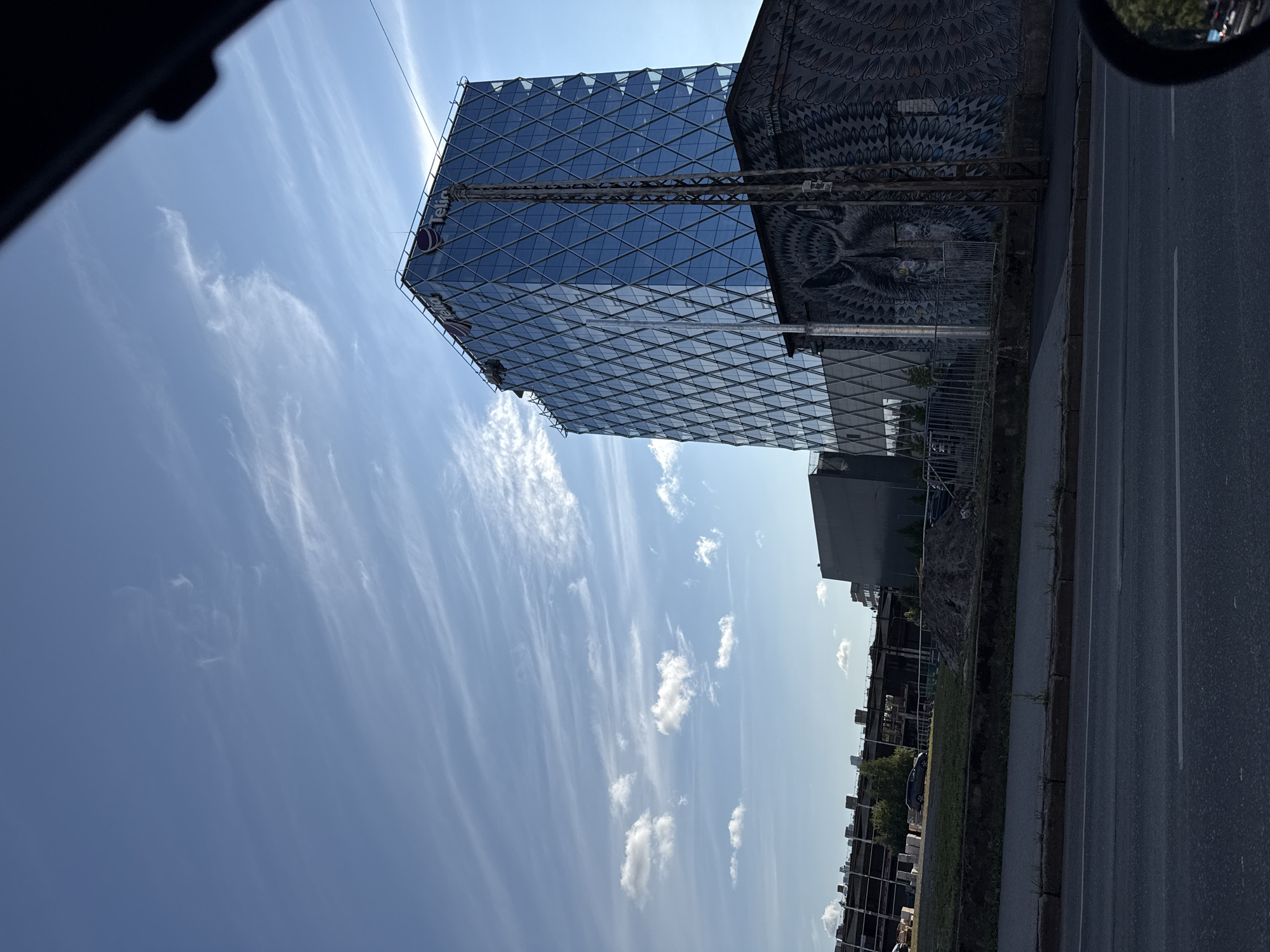 The image captures a bright, daytime scene in Tallinn, Estonia, featuring a modern glass-facade skyscraper with the "Telia" logo visible at the top. The skyscraper's diamond-patterned glass reflects the blue sky and wispy clouds. Adjacent to the skyscraper is a building adorned with a large mural of a wolf's face, rendered in a gray, black, and white palette, with feathered patterns on either side. In the background, a partially constructed building with visible concrete frameworks suggests ongoing urban development. The foreground shows a paved road with lane markings, a sidewalk, and a grassy embankment. The perspective suggests the photograph was taken from a vehicle, as indicated by the reflection in a car's side mirror in the lower right corner, showing streetlights and trees. The overall atmosphere is urban and contemporary, with a blend of new architecture and street art.