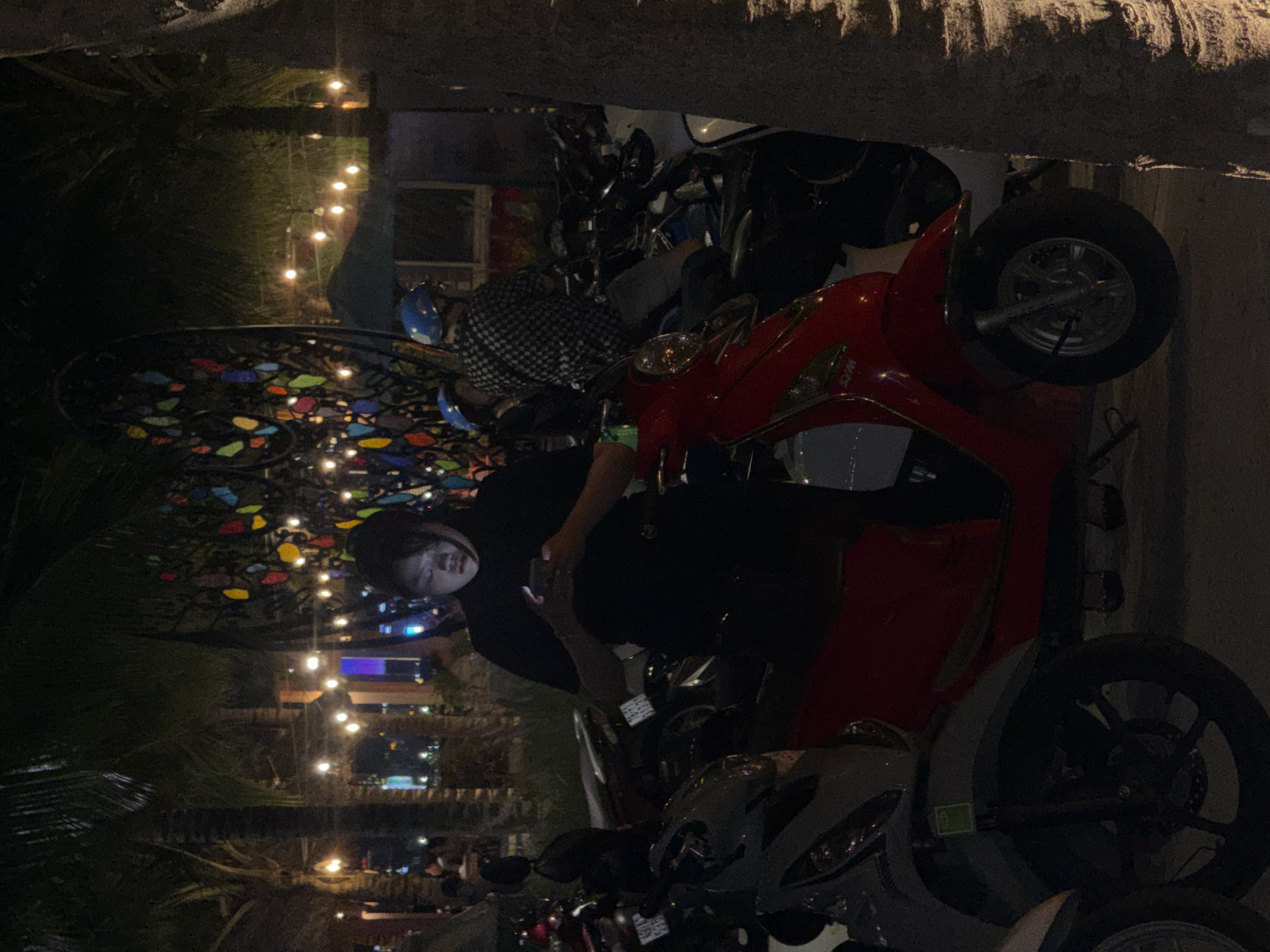 The image depicts an outdoor scene at night in what appears to be a public space in Bac Nha Trang Ward, Vietnam, likely a promenade or park area. Several parked scooters are visible, with a red scooter prominently in the foreground. A young person wearing a black long-sleeved shirt is standing next to the red scooter, looking down at a mobile phone they are holding. Behind this person, another individual is seated on a scooter, wearing a blue helmet and a black and white checkered shirt. In the background, there is a large, decorative, heart-shaped structure adorned with colorful glass pieces. String lights are strung across the area, illuminating the scene. Palm trees are also visible, indicating a tropical or subtropical environment. The overall atmosphere is dimly lit, with artificial lights contributing to the ambiance.