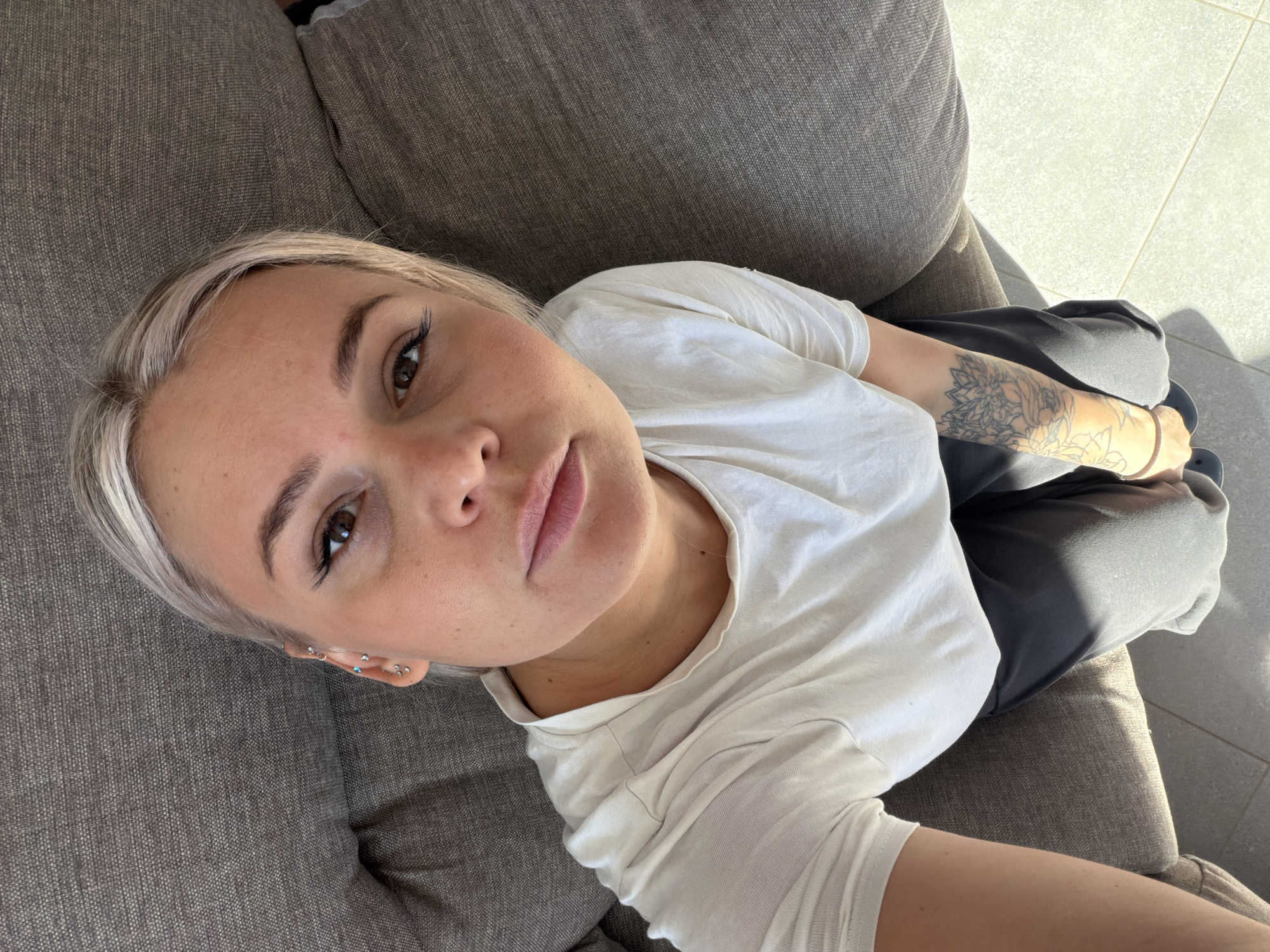 A young woman with short, light blonde hair is lying on a gray sofa. She is wearing a white t-shirt and dark-colored pants. Her right arm is visible, showing a floral tattoo. She is looking directly at the camera with a neutral expression. Multiple piercings are visible on her left ear. The background includes the sofa and a light-colored floor near a window, suggesting an indoor setting. There is no indication of the location being in Sarapul, Russia.