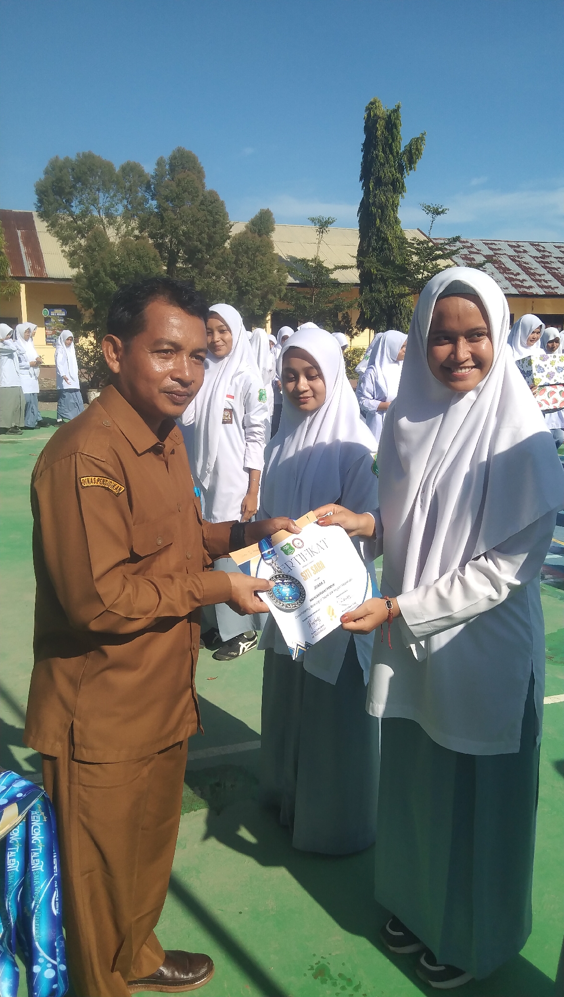 In Rhieng Blang, Indonesia, a man in a brown uniform, possibly a teacher or school official, presents a certificate and a medal to a smiling female student wearing a white hijab and uniform. The student is part of a group of other students in similar attire, suggesting an award ceremony or school event. The background shows school buildings with corrugated metal roofs and trees under a clear blue sky. The ground is a green surface, likely a sports field or courtyard. The overall scene appears to be an outdoor school setting during daylight hours.