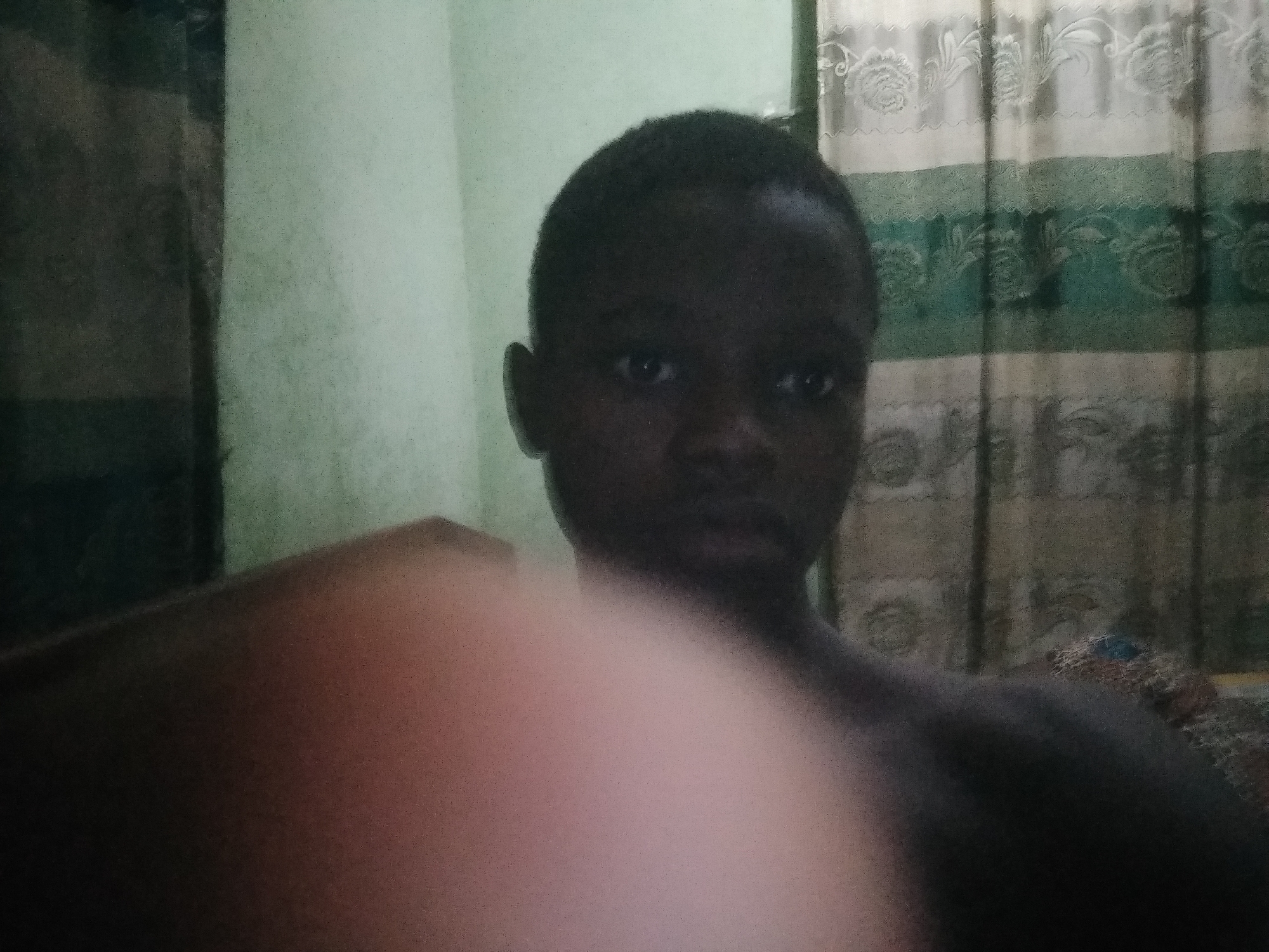 A person with dark skin is centered in the frame, their face visible, with a blurred object in the foreground obstructing part of the view. The background includes a wall painted in a light, greenish-gray color and curtains with a floral pattern in shades of white, green, and brown. The location is Unknown, Unknown.