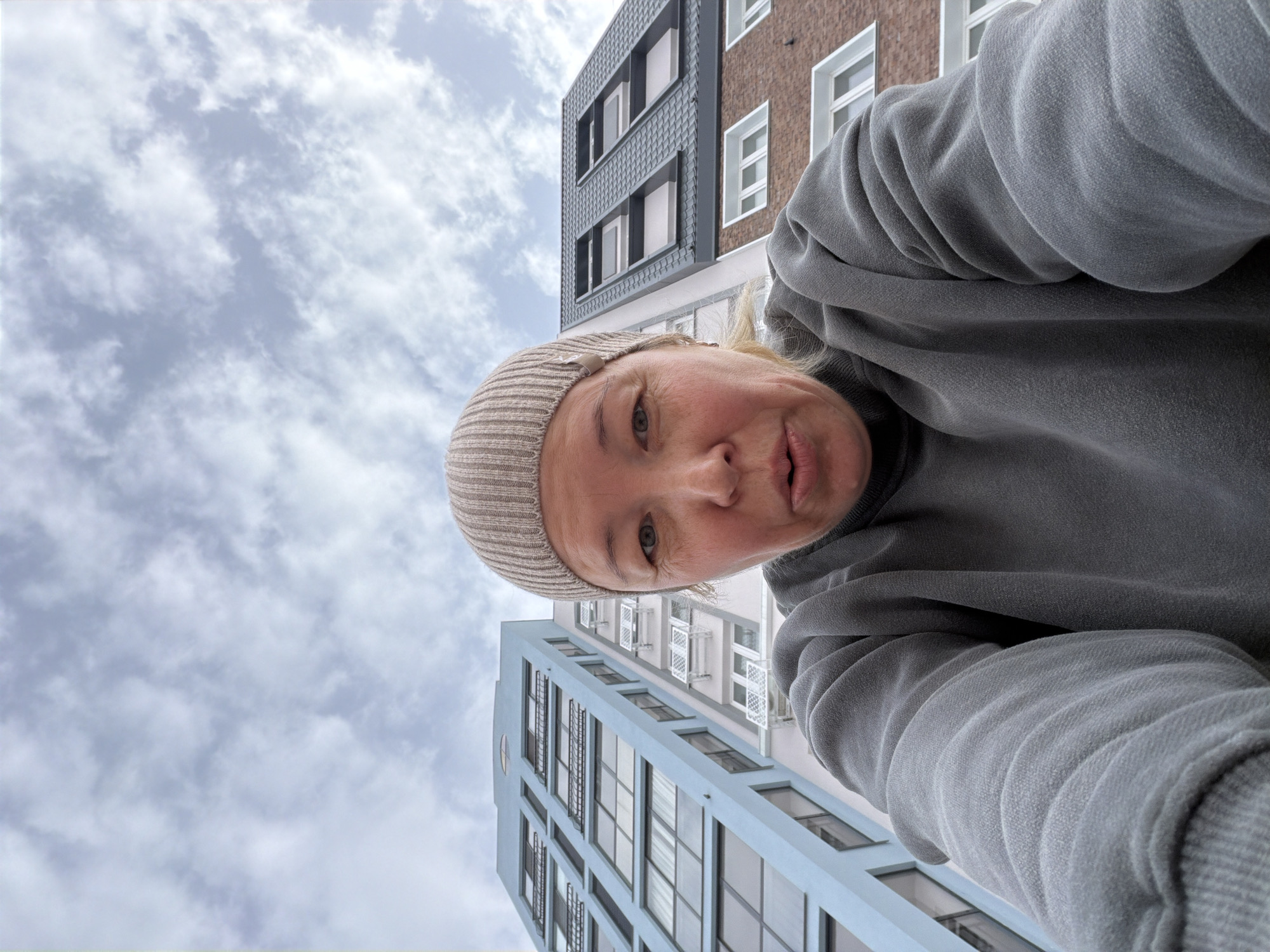 A woman in a tan beanie and gray sweatshirt is depicted from a low-angle perspective. Two buildings frame the background. The building on the left is light blue with multiple windows and dark accents. The building on the right is brick with white-framed windows and a gray trim. The sky is filled with scattered clouds. This image was probably taken in the city of Pervomaysky, Russia.