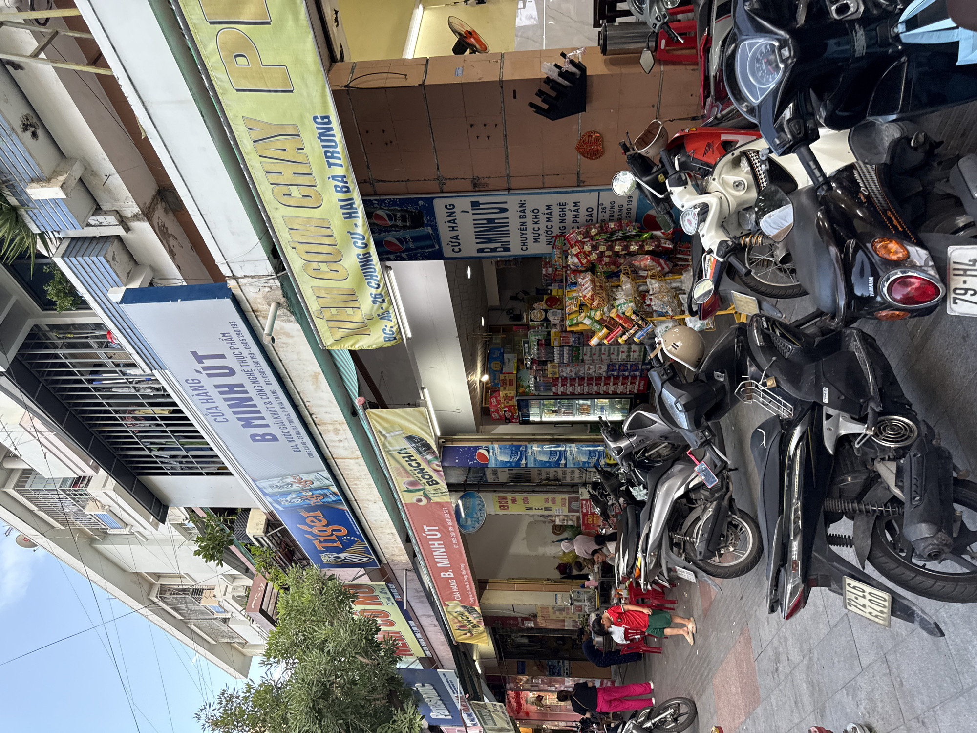 The image shows a street scene in Nha Trang, Vietnam, featuring a row of shops and parked motorcycles. A prominent shop sign reads "CỬA HÀNG B. MINH ÚT" (B. Minh Ut Store), indicating it sells food and beverages, with other signs mentioning "Tiger" beer and "Boncha". There are several motorcycles parked on the sidewalk and street, with some people in the background appearing to be customers or staff. The environment is urban, with buildings, overhead wires, and a blue sky visible. The overall impression is of a busy commercial area with typical street life.