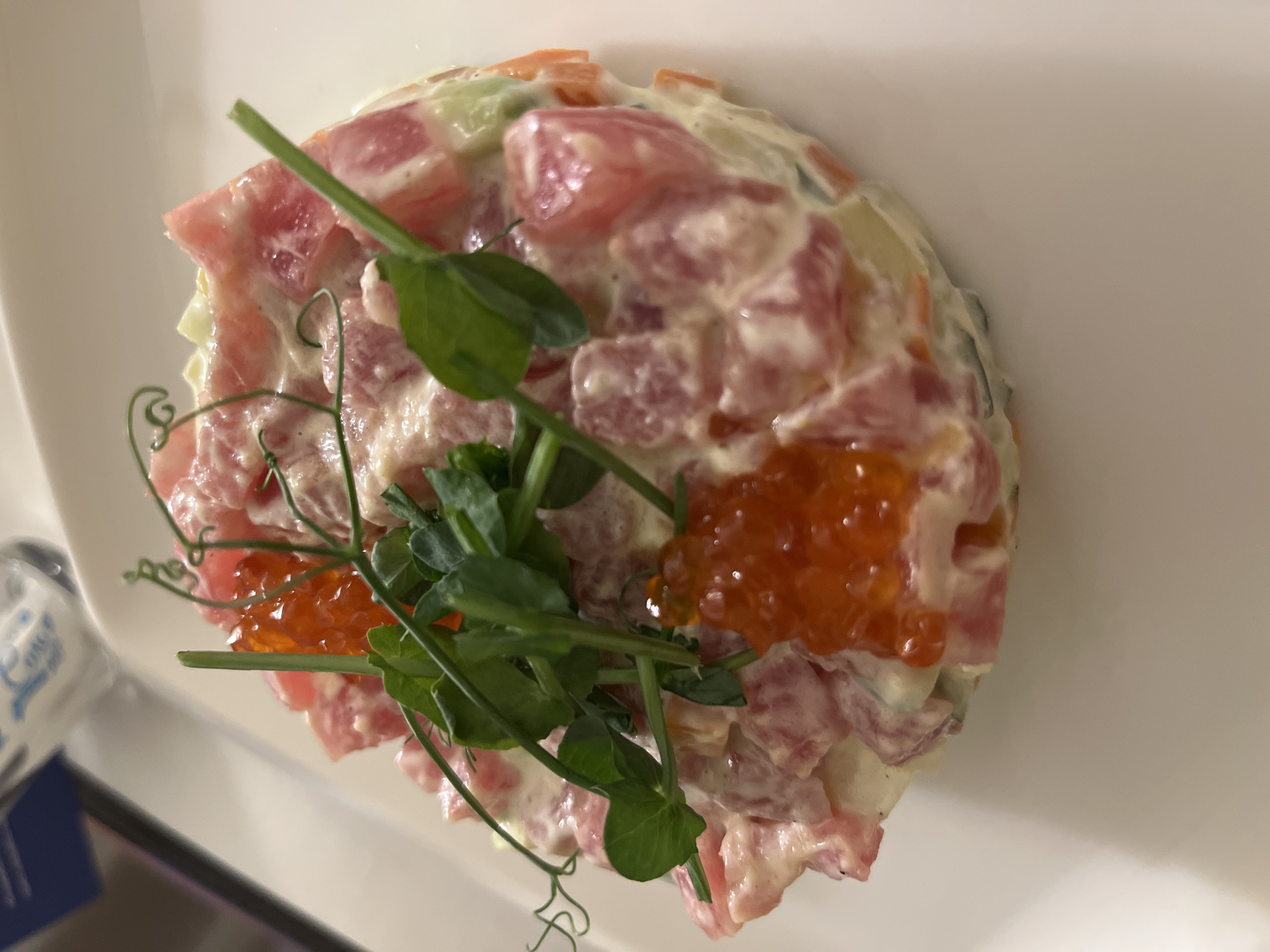A close-up, overhead shot shows a white plate on which is arranged a salad, featuring diced pink ingredients mixed with a creamy white dressing. The salad also includes orange-colored fish roe. Green pea shoots are placed on top of the salad as a garnish. The location and city cannot be determined from the image.