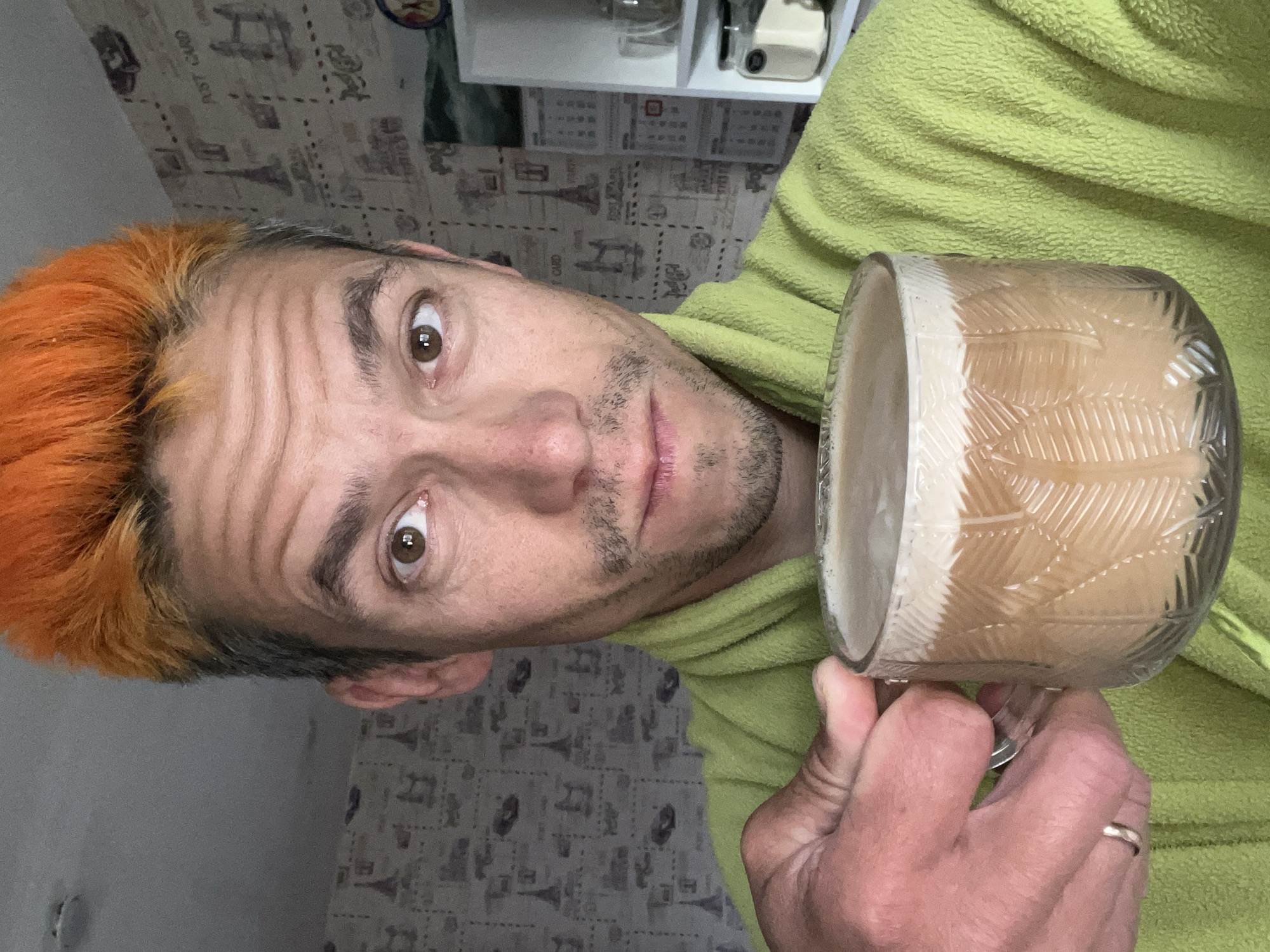 A selfie taken indoors shows a man with bright orange hair and a light green robe holding a mug of coffee. The background features patterned wallpaper with images of the Eiffel Tower and "Post Card" text. To the right, a white shelf unit is visible with various items, including jars and glassware. The man is looking directly at the camera. The scene appears to be a domestic interior, possibly a bedroom or living area, in Chaikovsky, Russia.