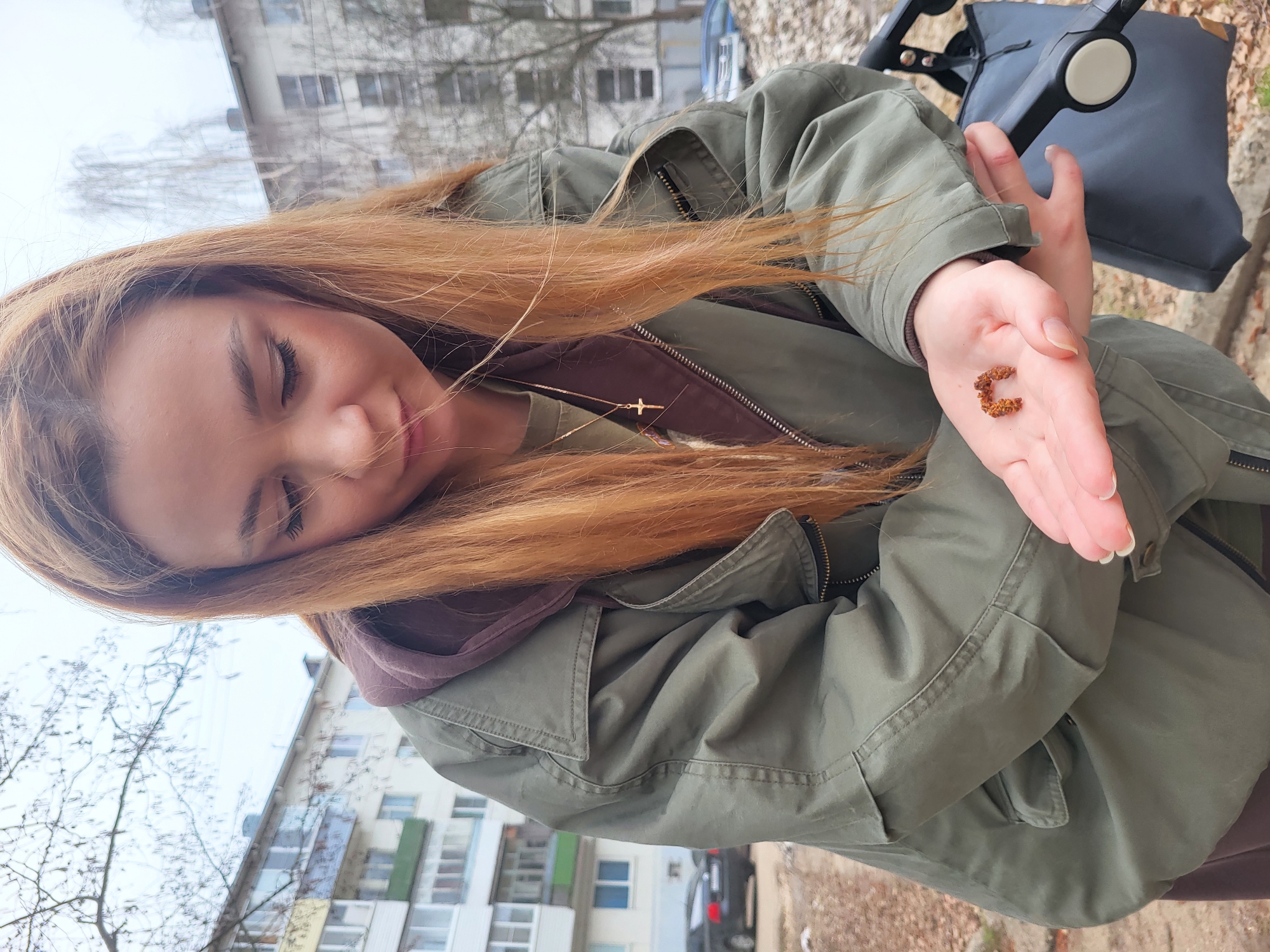 A woman with long, reddish-blonde hair is the central subject, photographed outdoors in what appears to be a residential area of Chaikovsky, Russia. She is wearing a green utility-style jacket over a dark brown hooded top and a delicate gold cross necklace. Her eyes are gently closed or nearly closed as she looks down at a small, reddish-brown, C-shaped object held in her open left palm. The object appears organic, possibly a seed pod, dried bark, or a piece of plant matter.
Behind her, a dark gray stroller or pram is partially visible, suggesting she may be with a child. The background features tall, multi-story apartment buildings, typical of urban or suburban environments, and bare trees, indicating a colder season such as late autumn, winter, or early spring. The sky is overcast, providing soft, diffused lighting. A dark car is visible parked in the background on the right side.
The woman's serene expression, with closed eyes, conveys a sense of calm and contemplation as she observes the object in her hand. Her fingernails are neatly manicured. No visible text is discernible in the image.