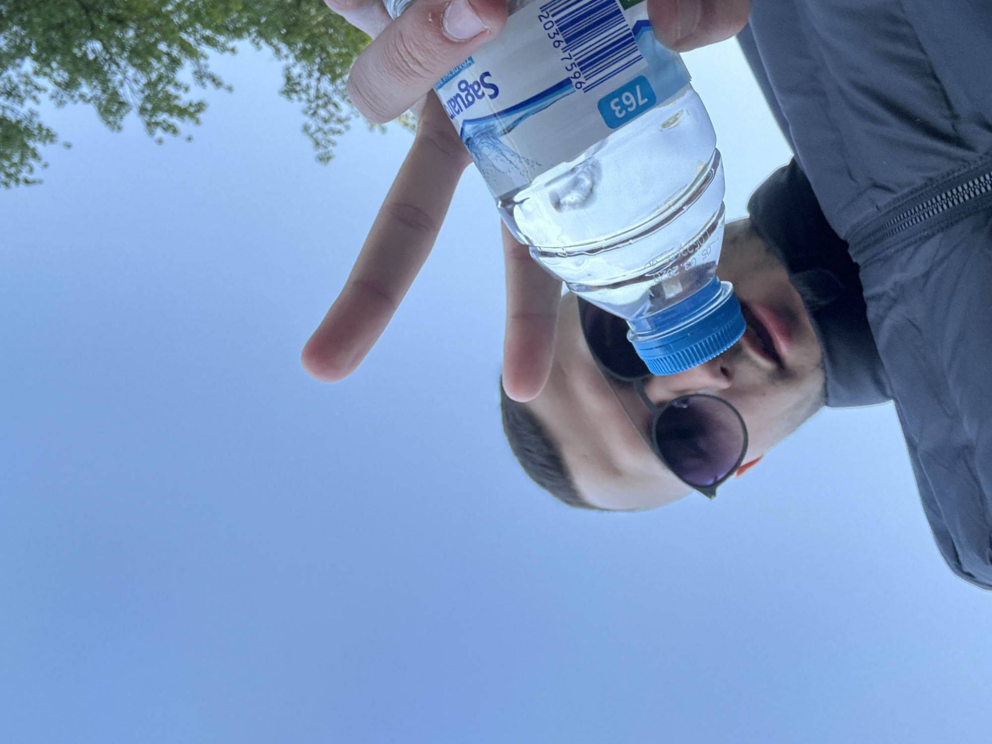 A low-angle shot looking up shows a man holding a small plastic water bottle with the "Saguar" brand visible. The man is wearing sunglasses and a dark jacket. Part of the sky and some tree leaves are visible in the background. There is no information in the image to suggest that this was taken in Plovdiv, Bulgaria.
