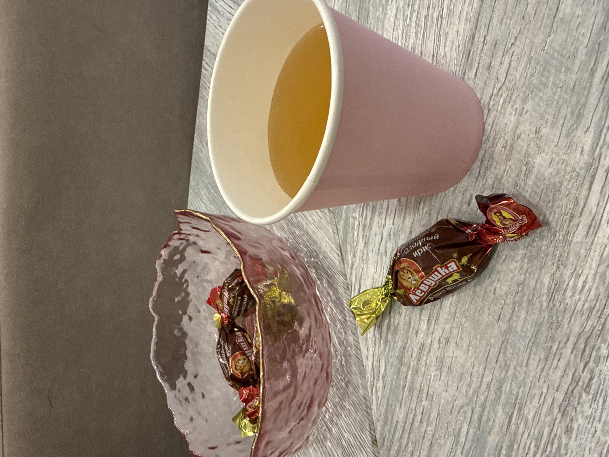 The image shows a pink disposable cup filled with a yellow liquid, a pink glass bowl filled with wrapped candies, and a single wrapped candy next to the bowl. The candies are labeled "Лёвушка" (Lyovushka), which translates to "Little Lion" and are produced by the SLADKO company. The scene is set on a gray, textured surface, likely a tabletop or counter. A portion of a gray sofa or chair is visible on the left side of the frame. Given the candy brand, the location is likely Saint Petersburg, Russia, where SLADKO is based.