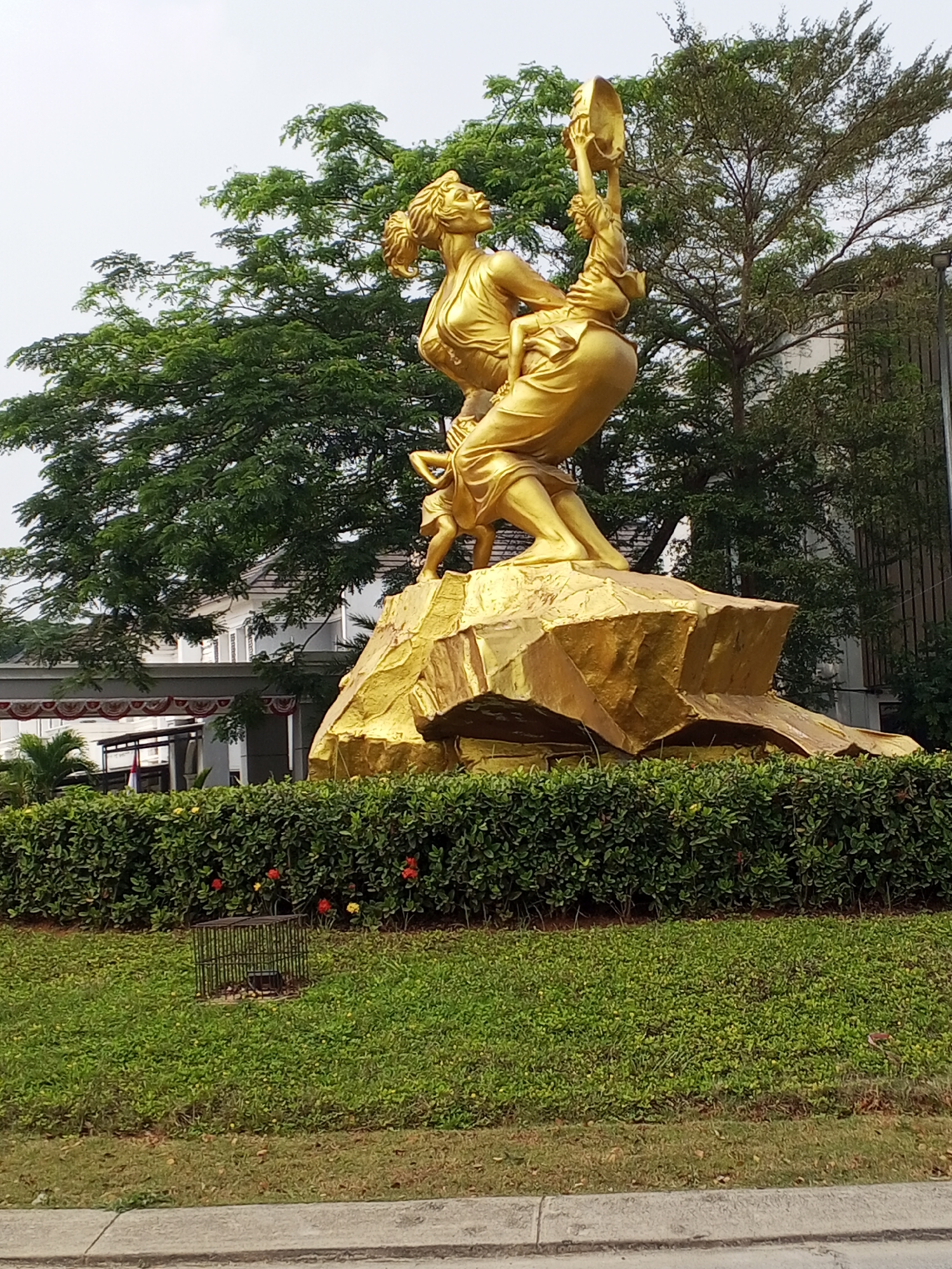 A golden statue of a woman holding a round object above her head is prominently featured in the foreground of the image. She is positioned on a rocky, golden base, which is surrounded by a dense, green hedge. Behind the hedge, a well-maintained lawn extends, with a small, caged structure visible on the grass. In the background, lush green trees and the facade of a building are present. The sky is overcast. This statue is located at the Taman Kota Serang, Serang, Indonesia.