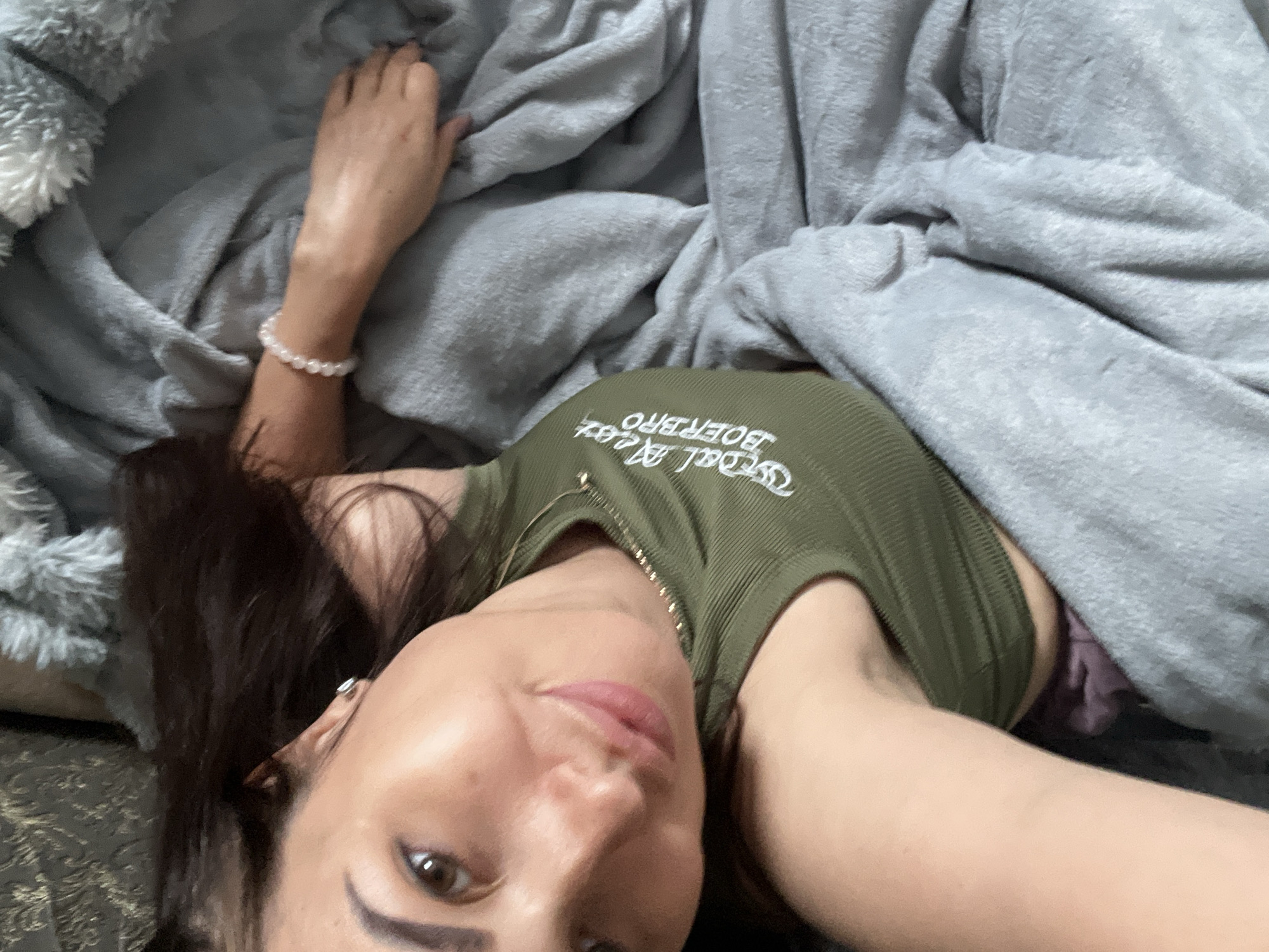 The image shows a person lying down on a bed covered by a grey blanket. The person's face is turned towards the camera, and they are wearing a green tank top with white text on it. The text appears to read "WEDAL AT BOERBRO". The person has dark hair and is wearing a bracelet on their left wrist and a small silver earring in their left ear. The lighting suggests it could be daytime, but the exact time of day and location are unknown. The scene appears to be casual and intimate, possibly taken in a bedroom.