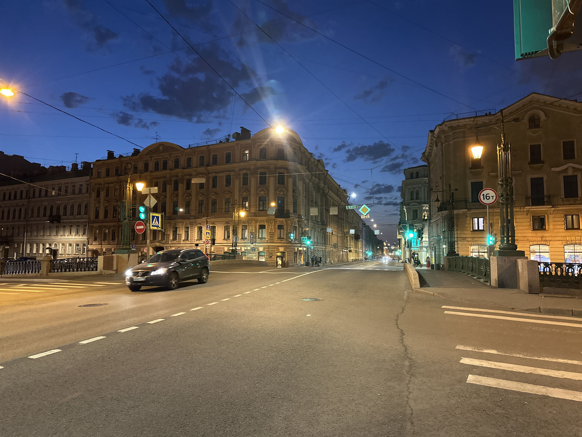 This image shows an urban scene in Saint Petersburg, Russia, at dusk or dawn. The perspective is from a road on a bridge, looking down a straight street. A dark-colored SUV is driving along the road toward the viewer, its headlights illuminated. The street is lined with buildings of historical architecture, illuminated by street lamps and lights from within. Several traffic lights are visible along the street, all showing green. Pedestrian crossings are marked on the road. People are sparsely seen walking on the sidewalks. The sky is a mix of blue and dark cloud cover. Wires run across the top of the image, possibly for electrical or tram lines. Street signs with Cyrillic lettering are visible, along with a road sign with a 16t marker.