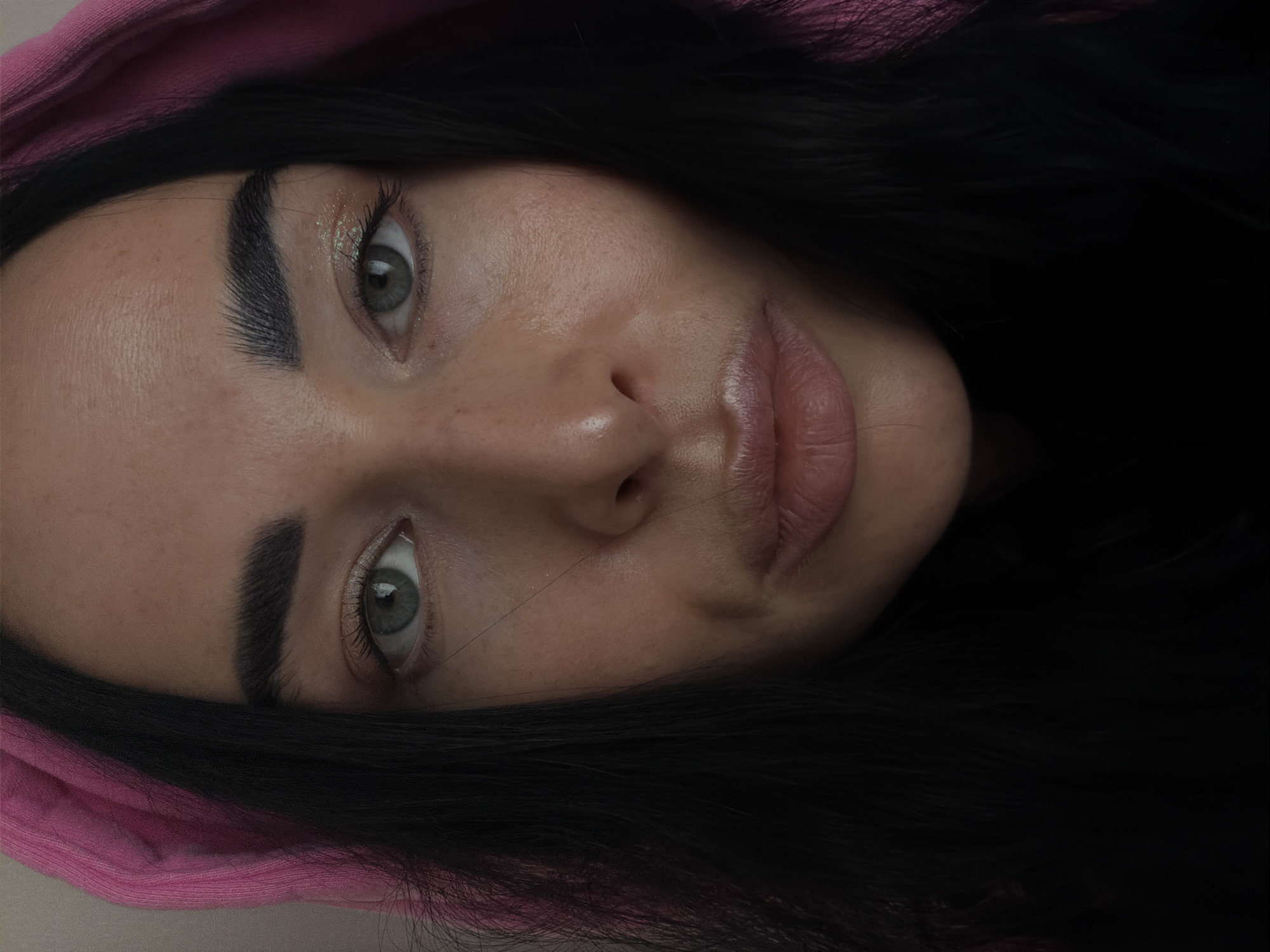 A close-up, portrait-style photograph depicts a woman's face, slightly tilted. She has fair skin, dark hair, and light-colored eyes. The woman is wearing a pink hooded garment and has dark eyebrows. Traces of glittery eye makeup are visible. There is no discernible indication that the image was taken in Saint Petersburg, Russia.