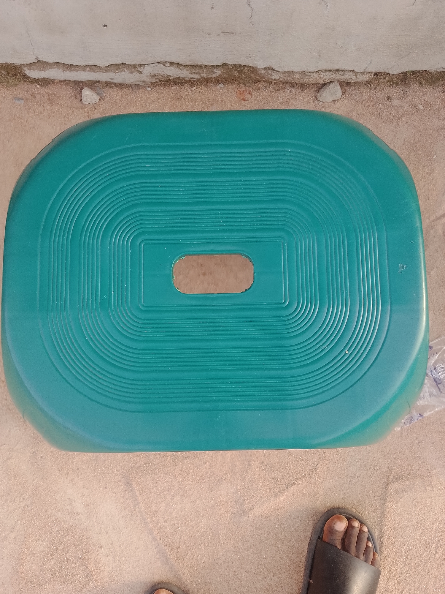 An overhead shot shows a teal plastic stool placed on a light brown sandy surface. The stool is rectangular with rounded corners and features concentric ridges on its surface, forming a patterned top. A rectangular cutout is visible in the center of the stool. In the lower right corner of the image, a bare foot wearing a black sandal is visible. The background consists of a light-colored wall with a rough texture, and a strip of pebbles and dirt separates the wall from the sandy ground. The overall scene appears to be outdoors.
