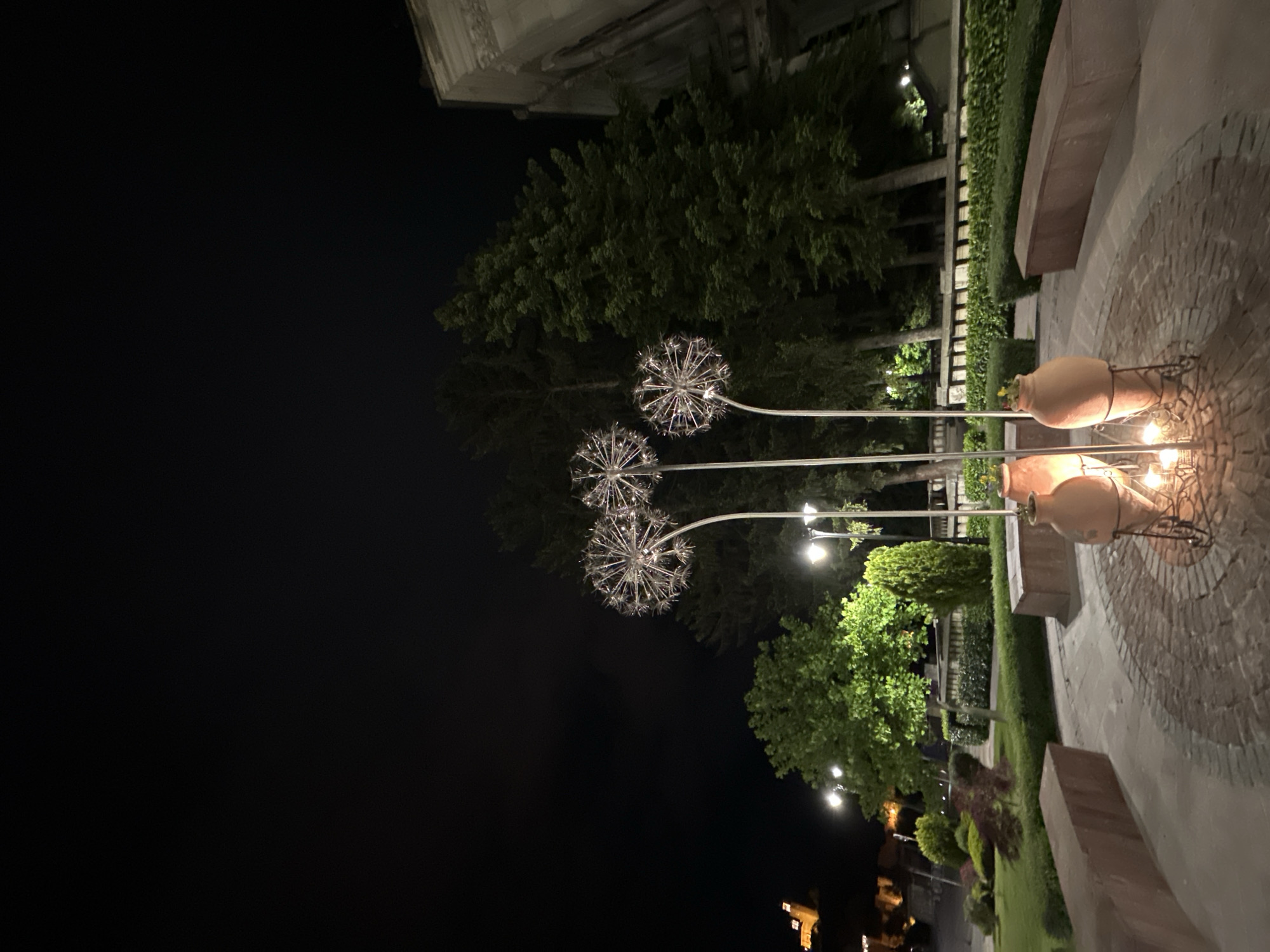 At night, the city park in Veliko Tarnovo, Bulgaria, presents a peaceful scene. Ornamental light fixtures, resembling dandelions, dot the landscape. Adjacent to these are three clay pots on stands, each lit with a warm glow, potentially housing candles or other light sources. The park's perimeter is defined by stone benches, bordering a meticulously maintained hedge. Across the landscape, a variety of trees and shrubbery are illuminated by streetlights, and further in the distance, city lights twinkle.