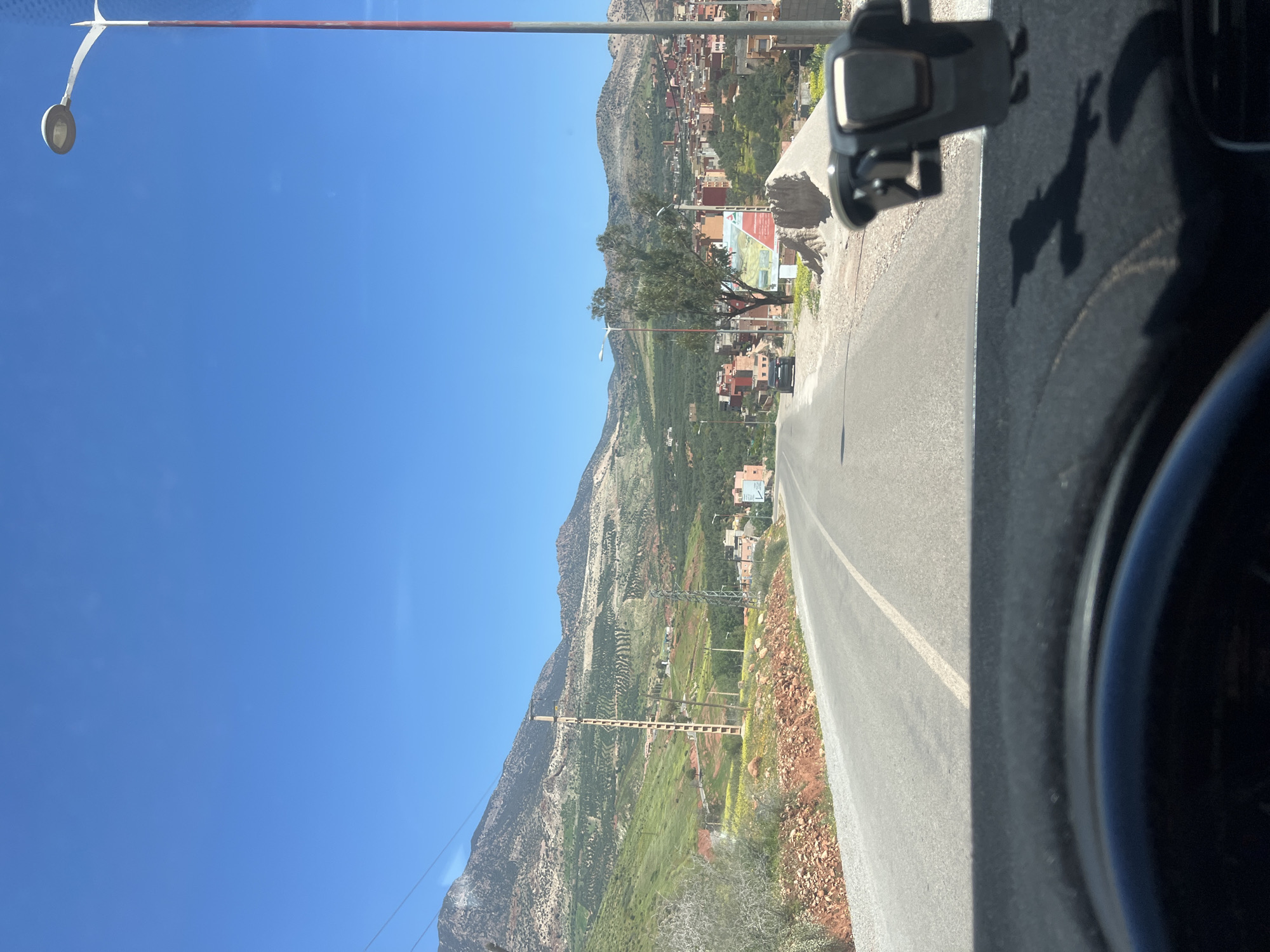 The image shows a view from inside a vehicle traveling on a road near the city of Ouaouizert, Morocco. The landscape is mountainous, with a mix of green vegetation and rocky terrain. Houses are scattered along the hillsides, typical of settlements in mountainous regions. An electrical pylon runs alongside the road. The sky is clear blue. A street light is visible on the left. The dashboard of the vehicle is partially visible on the right, including a phone mount and a shadow. A sign can be seen along the road.