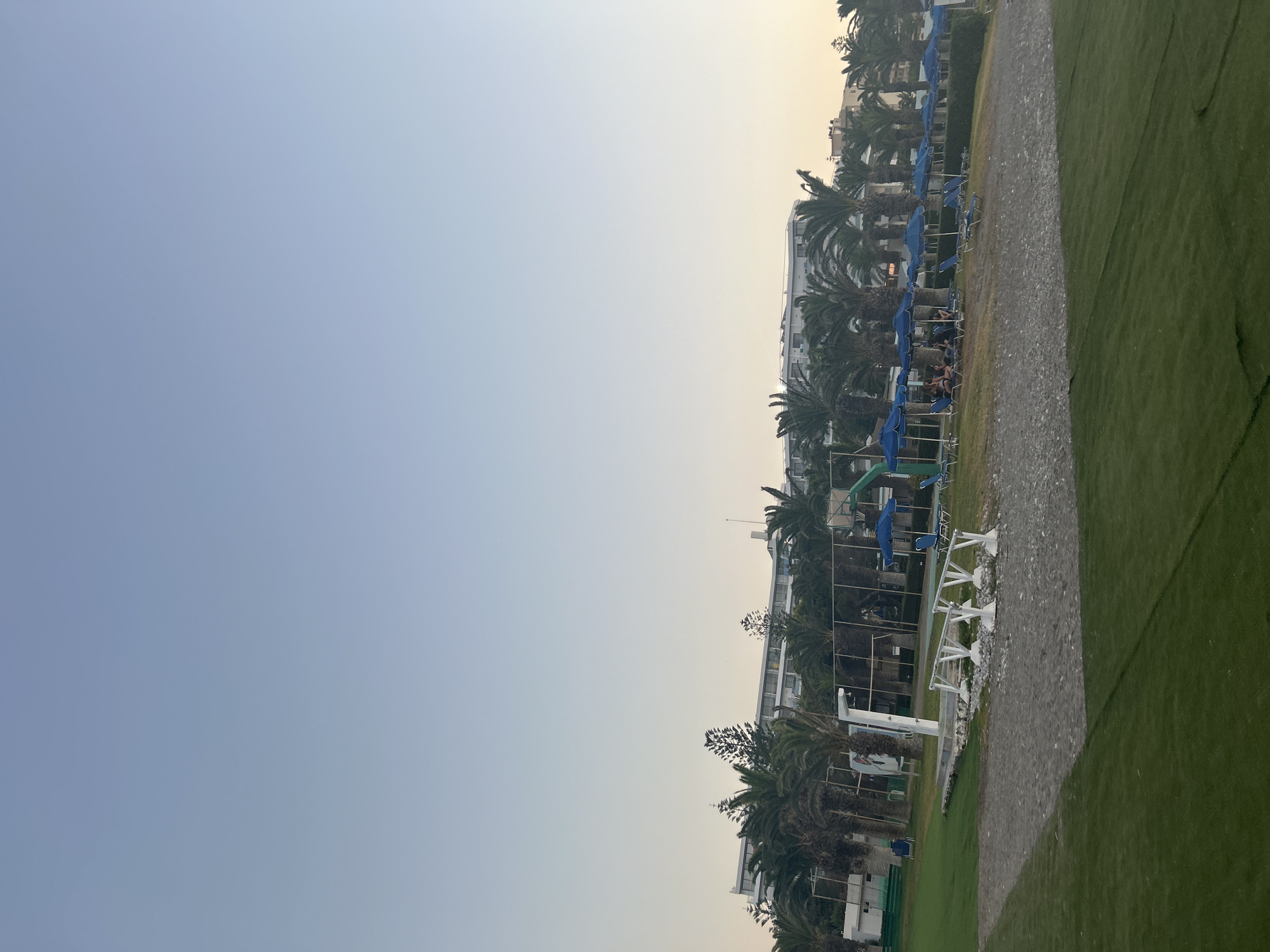 This media file shows a view of a resort or hotel area with a lawn, palm trees, and buildings. The scene is dominated by a clear, pale blue sky that transitions to a hazy yellow or white near the horizon, suggesting either dawn or dusk. In the mid-ground, there is a row of buildings with white facades and visible windows. In front of the buildings, a line of blue umbrellas and lounge chairs are set up on a gravel or sand path. Several palm trees are scattered throughout this area, casting shadows. The foreground features a strip of green grass. There appear to be a few people in the lounge chairs, some of whom are wearing swimwear, suggesting a recreational setting like a beach resort or a pool area. The overall atmosphere is calm and relaxed. The image is oriented vertically. No visible text is present. The location is identified as Rhodes, Greece.